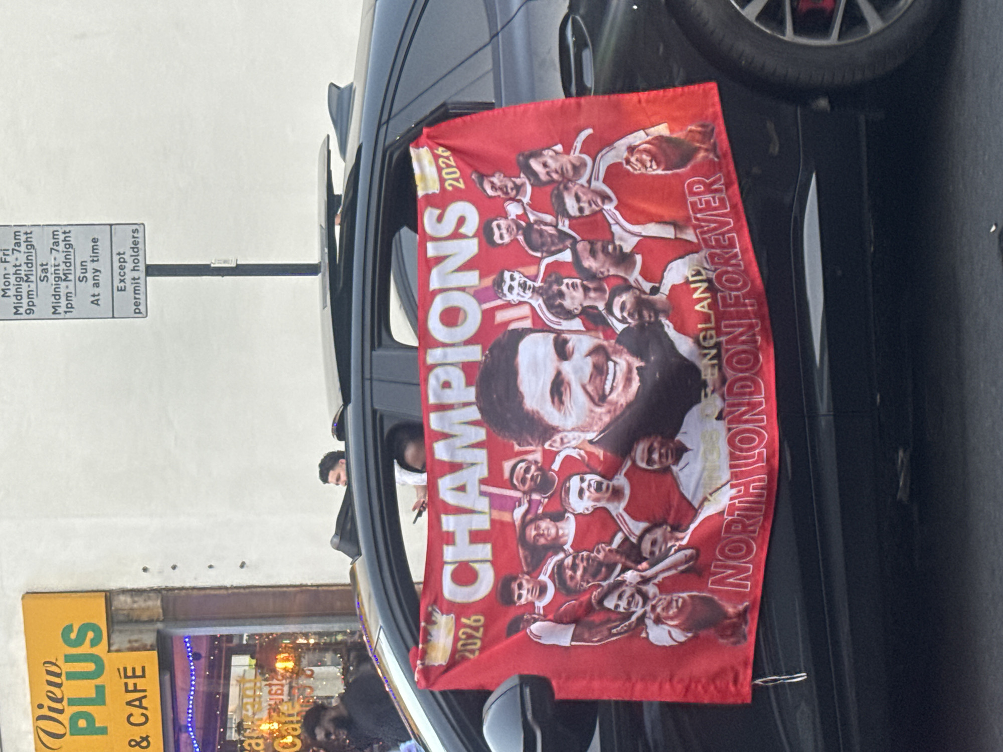 The image shows a black car parked on a street in London, United Kingdom. The car has a red flag hanging from the driver's side window. The flag features an image of a group of men, presumably a sports team, with the words "CHAMPIONS 2026" and "NORTH LONDON FOREVER."
In the background, there's a yellow sign reading "View PLUS & CAFÉ" with a cafe's interior visible through the windows. A parking sign is also visible that provides information on restricted parking, except for permit holders, at different times of the day and week. Pedestrians are partially visible near the cafe.