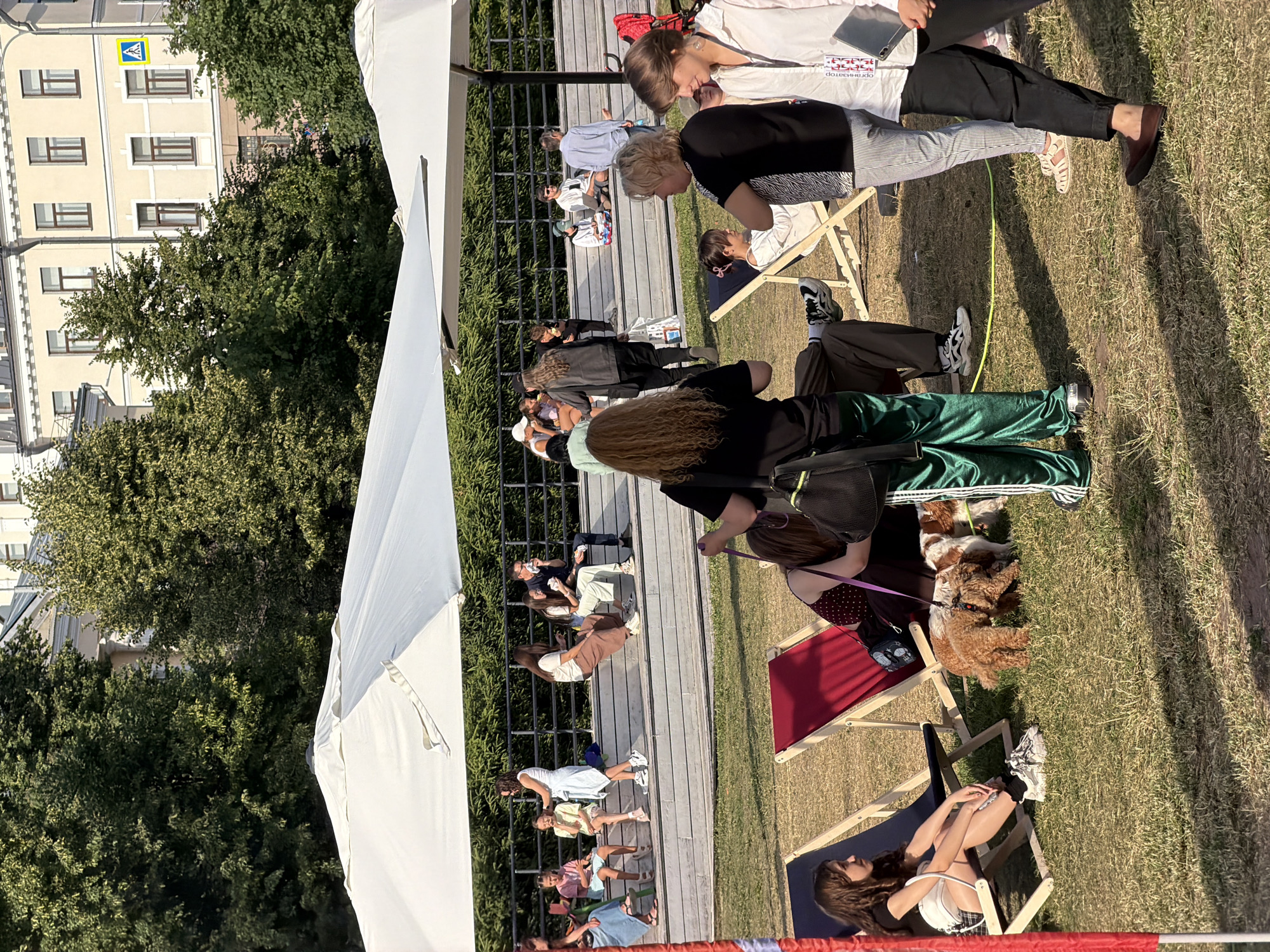 This media file captures an outdoor gathering in Kazan, Russia, under what appears to be pleasant weather with a bright sun casting shadows. The scene is set on a grassy area with a wooden walkway and tiered seating.
Several groups of people are present, mostly young adults and some children, engaged in a relaxed, social event. Some individuals are seated on the tiered wooden steps, while others recline on portable lounge chairs. The atmosphere seems informal and casual, suggesting a picnic, outdoor concert, or similar community event.
Notable details include two dogs in the foreground, one a light brown cockapoo and another a darker brown, both appearing relaxed on the grass. A large white tent or canopy structure is visible on the left side of the frame, possibly providing shade. In the background, a multi-story building with numerous windows can be seen, along with lush green trees. The time of day is likely afternoon, given the angle of the sun and the long shadows.
One of the individuals in the foreground, seated on a lounge chair, is wearing a white top with what appears to be text in Russian. The legible portion appears to read "организатор" which translates to "organizer". This suggests that some individuals might be involved in the planning or execution of the event. The overall mood is one of leisure and enjoyment, with people conversing, observing, or simply taking in the surroundings.