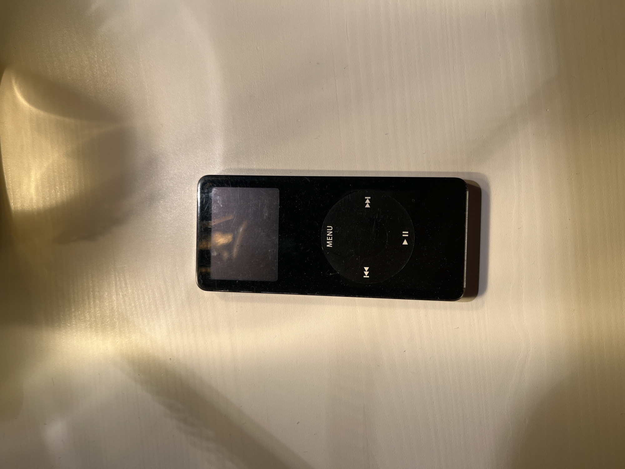 A black iPod Classic is centered on a light-colored wooden surface. The iPod is oriented vertically. Its screen is dark, reflecting some light. Below the screen is a circular click wheel with a play/pause button in the center, a rewind/skip backward button to the left, and a fast-forward/skip forward button to the right. The surface appears to be a table or desk, with textured wood grain visible. Above and to the left of the iPod, there are shadows and reflections of light that create a patterned effect on the surface, suggesting overhead lighting. The background is out of focus. The scene is indoors, likely in a home or office environment.