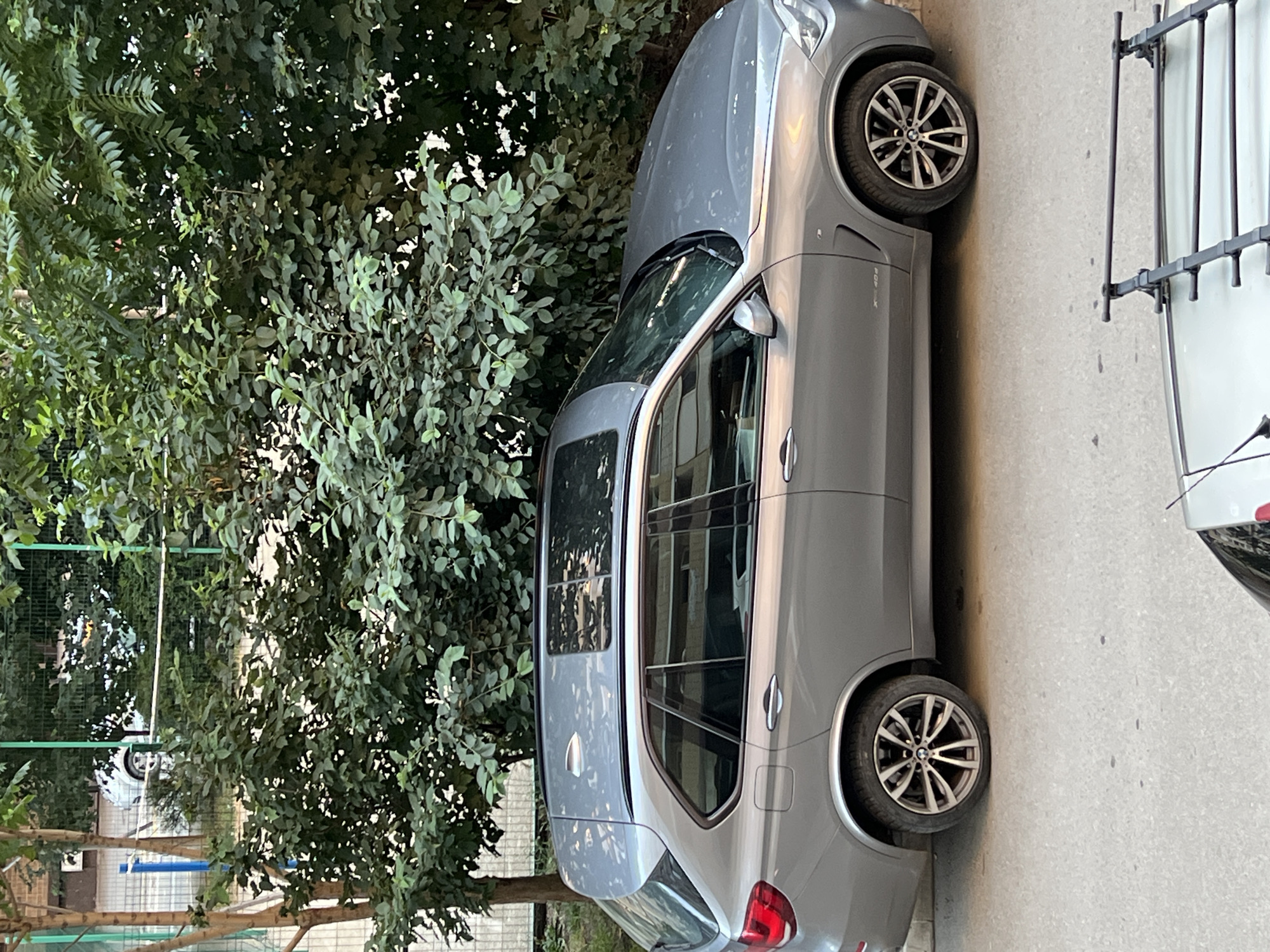 A silver BMW X5 SUV is parked on a paved street in Krasnodar, Russia. The vehicle is positioned parallel to the curb, facing right. Dense green trees and foliage are visible behind the car, casting shadows. In the foreground, the roof of another car with a roof rack is partially visible. The scene appears to be an outdoor urban environment, possibly a residential area.