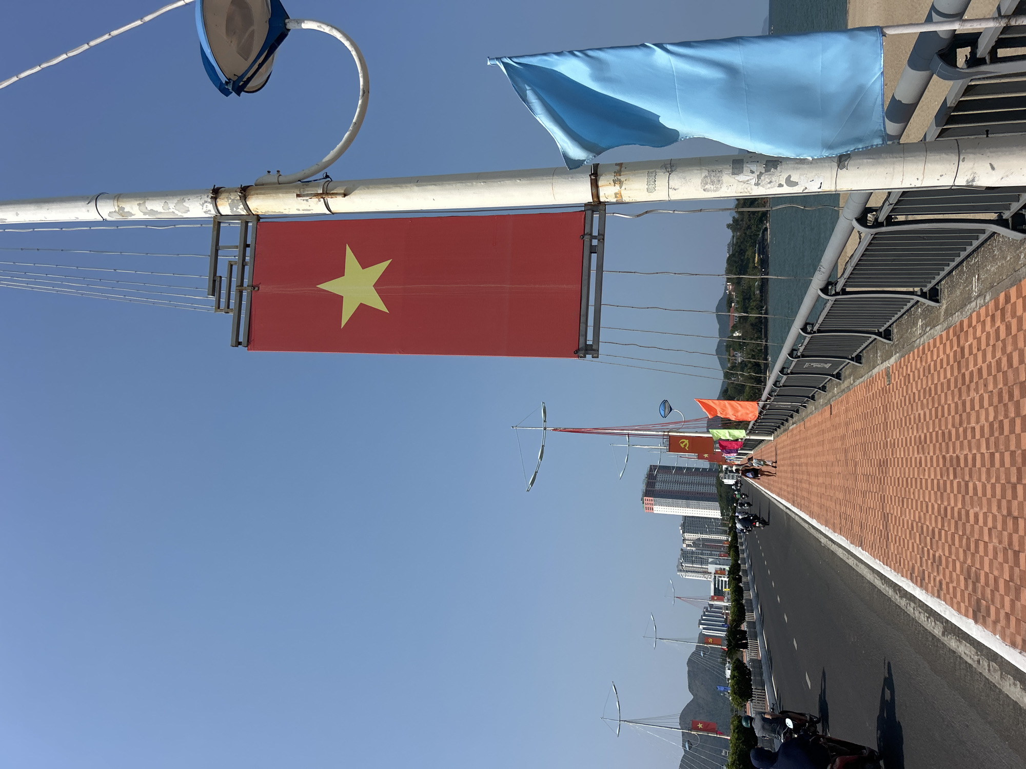 The image shows a view along a bridge in Nha Trang, Vietnam. A Vietnamese flag hangs from a street light on the left. Another blue flag is visible on the same post. The bridge features a brick-patterned walkway on the right, separated from the asphalt road by a white curb. Further down the bridge, other flags and light poles can be seen. Buildings rise in the background, with mountains visible in the distance. Traffic, mainly motorbikes, is using the bridge. The sky is clear and blue.