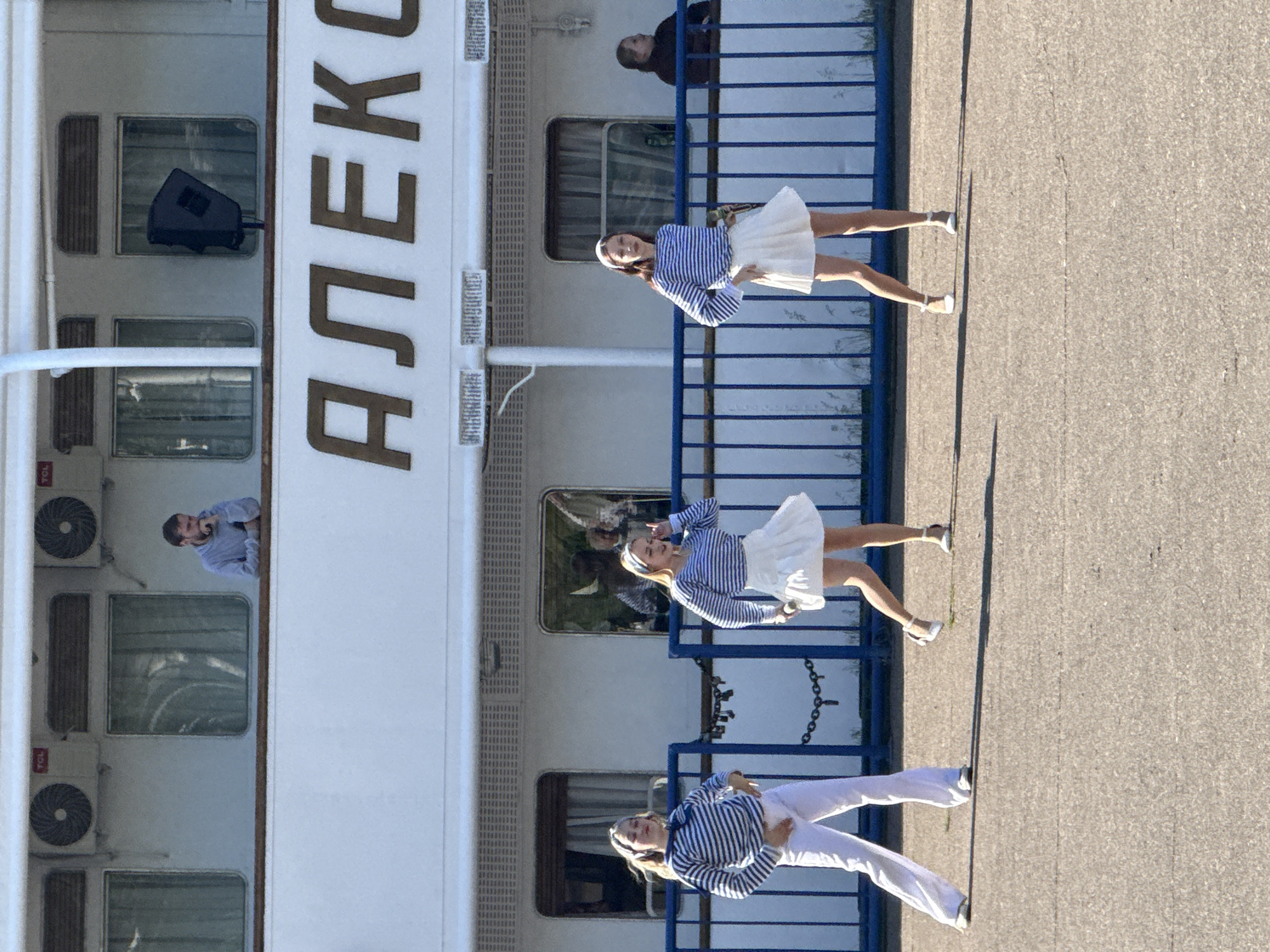 The image captures a scene on or near the ship 'АЛЕКСЕЙ', likely docked near the city of Chaikovsky, Russia. Three women are positioned near a blue railing, wearing nautical-themed outfits of blue and white striped tops, white skirts, and white shoes. They appear to be posing or performing. A man leans out of a window in the upper deck. Another person is on the deck. The background includes the ship's hull, windows, and air conditioning units. The location is paved.