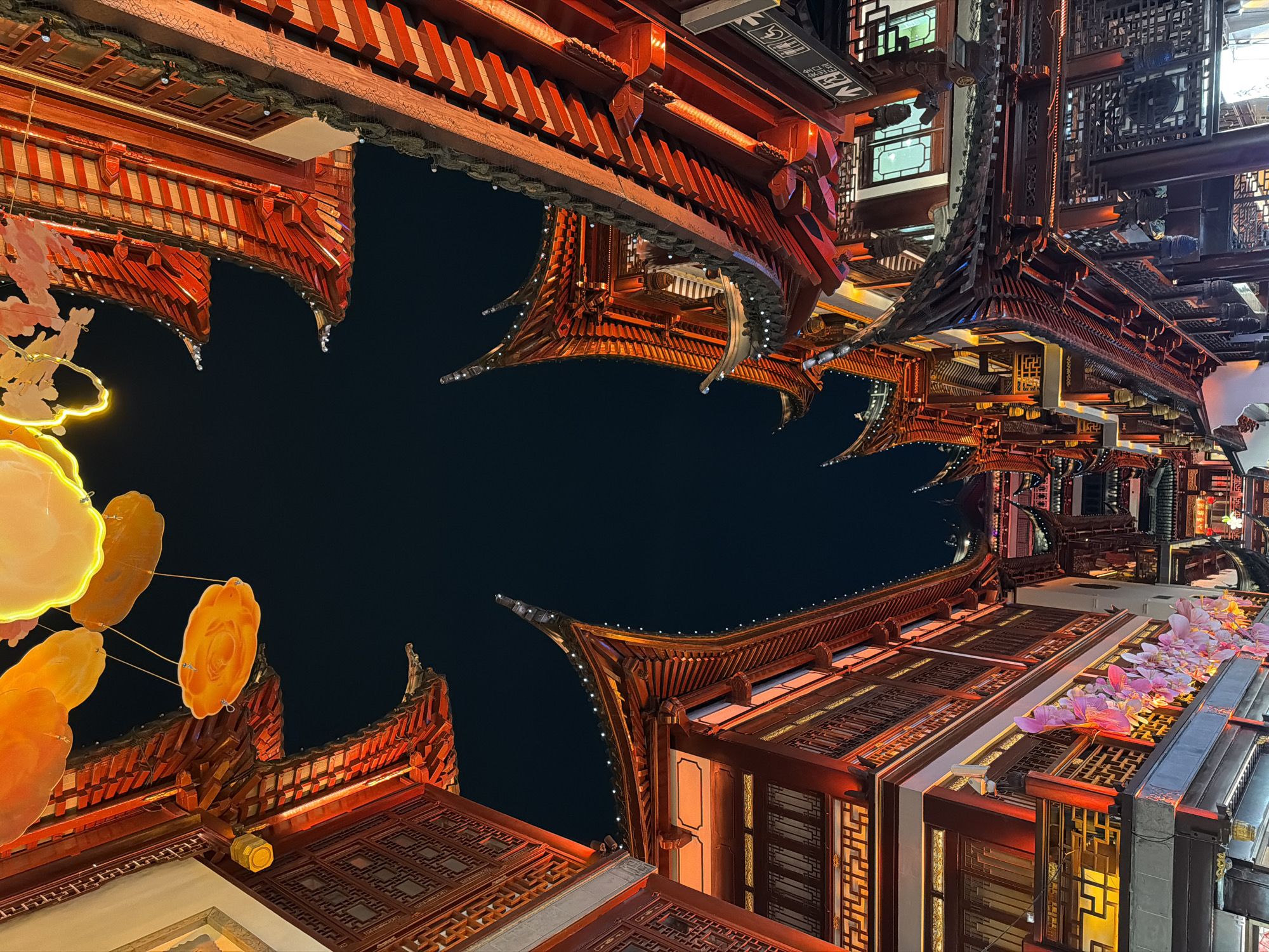 The image depicts a nighttime view looking up at traditional Chinese architecture in Shanghai, China, likely in the Yu Garden area. The buildings are adorned with intricate red and brown woodwork, and the eaves are curved. Decorative lights, including large flower-shaped lanterns, are present. The sky is dark. The image is vertically oriented with the viewer looking up.