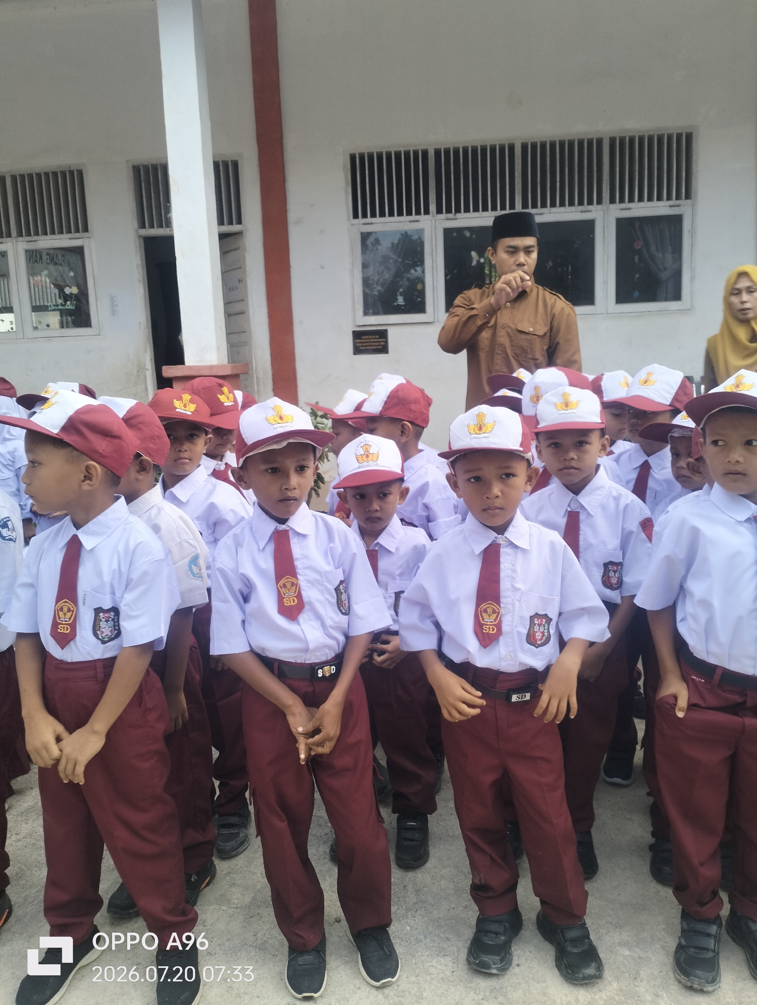 A group of young school children in uniform stand outside a building. The children are wearing white shirts with red ties and maroon pants. They also have on red and white hats with school emblems. Some of the children are looking at the camera, while others are looking away. In the background, an adult male wearing a brown shirt and a black cap, possibly a teacher or administrator, is standing. To the right of the male, a woman in a yellow headscarf is visible. The building appears to be a school with barred windows. The ground is paved with concrete. The overall scene suggests a school event or assembly.