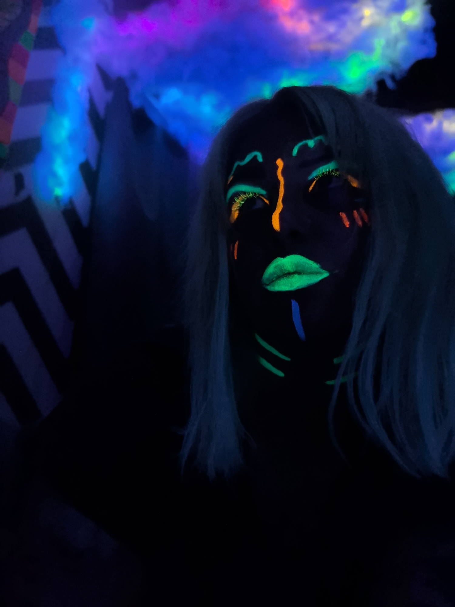 This striking image captures a person adorned with elaborate neon face paint under ultraviolet light, creating a vibrant and otherworldly effect. The central subject is seen from the chest up, with their face angled slightly upwards and to the right, against a dark background. Their skin appears dark under the UV light, emphasizing the glowing makeup.
Notable details of the face paint include bright neon green lipstick, neon green lines outlining the eyes, and distinct neon green/yellow eyelashes. A prominent orange line runs vertically down the bridge of the nose, extending onto the forehead. Short, glowing orange and red dashes are visible on the cheeks, while abstract neon green and blue lines decorate the chin and jawline. The person's hair, appearing light blue or silver under the UV illumination, frames their face.
The background is dark, featuring luminous, cloud-like formations in shades of blue, purple, pink, and green, suggesting an artistic lighting setup or fog effects. To the left, a distinct black and white chevron or zigzag striped pattern adds a graphic element to the scene. The overall impression is that of a performance, party, or photoshoot in a dark, indoor environment, likely at night, designed to maximize the impact of glow-in-the-dark materials. The image evokes a sense of creative expression and an energetic atmosphere. There is no visible text in the image. The location context is Noviy, Russia, though no specific visual cues within the image confirm this.