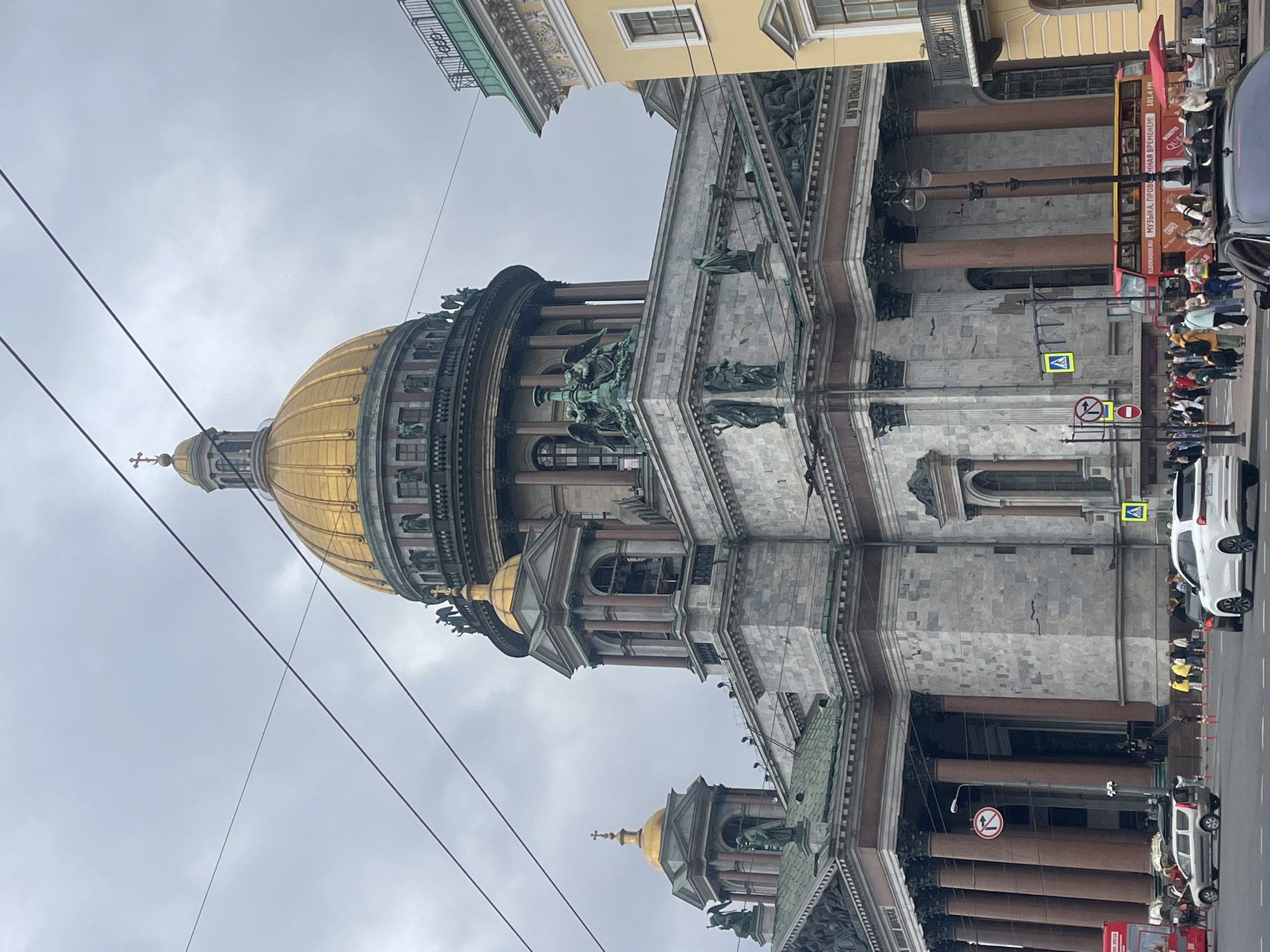The image shows a frontal view of Saint Isaac's Cathedral in Saint Petersburg, Russia, under a cloudy sky. The cathedral, a grand neoclassical building, features a prominent golden dome topped with a cross. The facade is characterized by large Corinthian columns, statues, and intricate architectural details. In the foreground, a street scene unfolds with several vehicles, including a white SUV, a black sedan, and a red double-decker bus, suggesting a public space or tourist area. Pedestrians are visible walking on the sidewalk and crossing the street. Traffic signals and street signs are present, indicating an urban environment. The presence of the double-decker bus, a common tourist vehicle, and the impressive architecture of the cathedral strongly suggest this is a popular tourist attraction within the city. The overall mood is subdued due to the overcast weather.