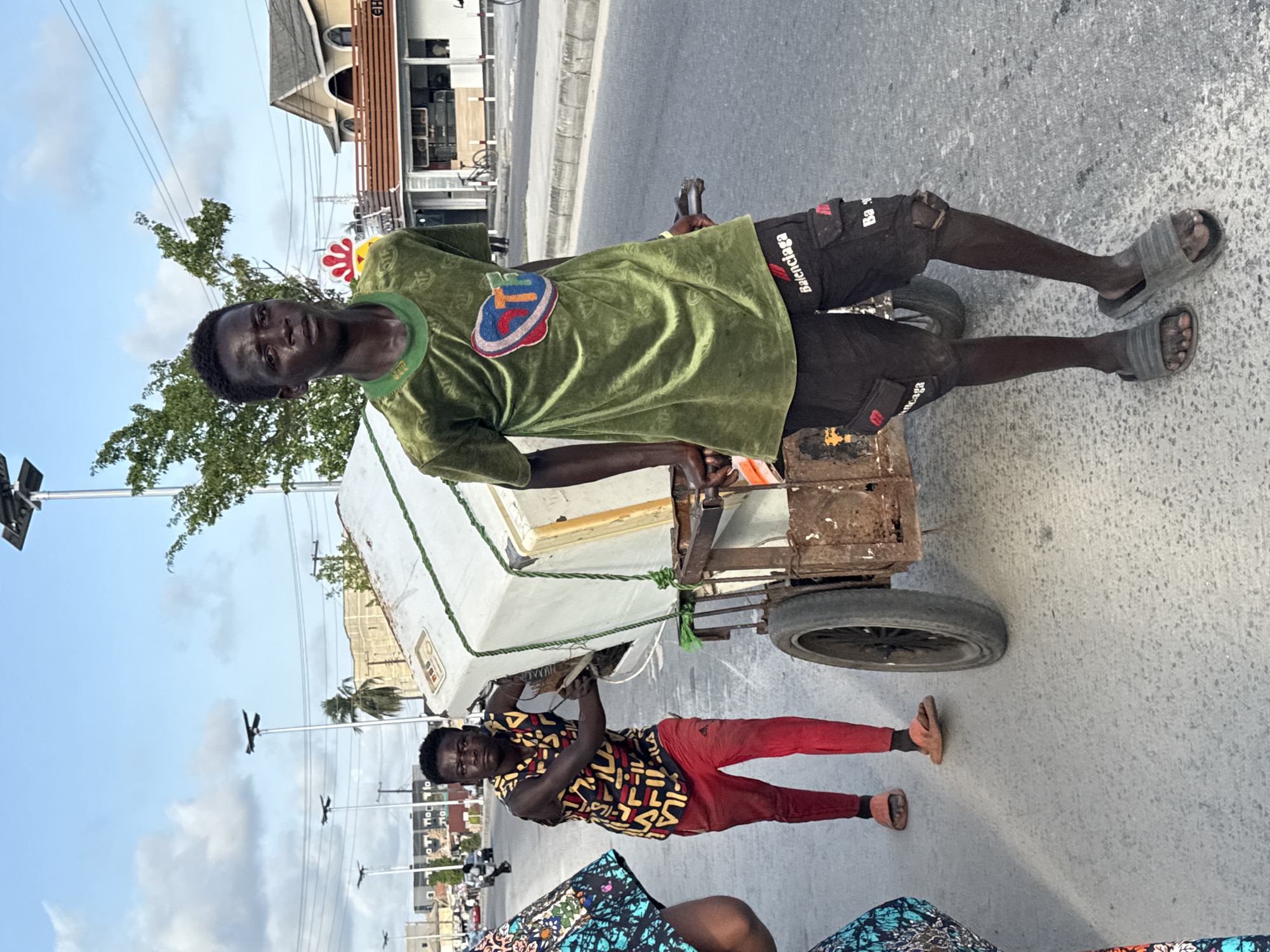 Two African men are on a street in Ado, Nigeria. The man on the left, in a green T-shirt and black shorts with "Balenciaga" in white lettering, is standing next to a white and silver cart with two wheels. The man is wearing grey slides. The cart is rusted and appears to be carrying something under a white cover. The man on the right is partially holding the cart and is wearing a patterned black and yellow shirt with red pants and brown slides. The street is grey and there are buildings and trees in the background. There are overhead electrical wires. The sky is blue.