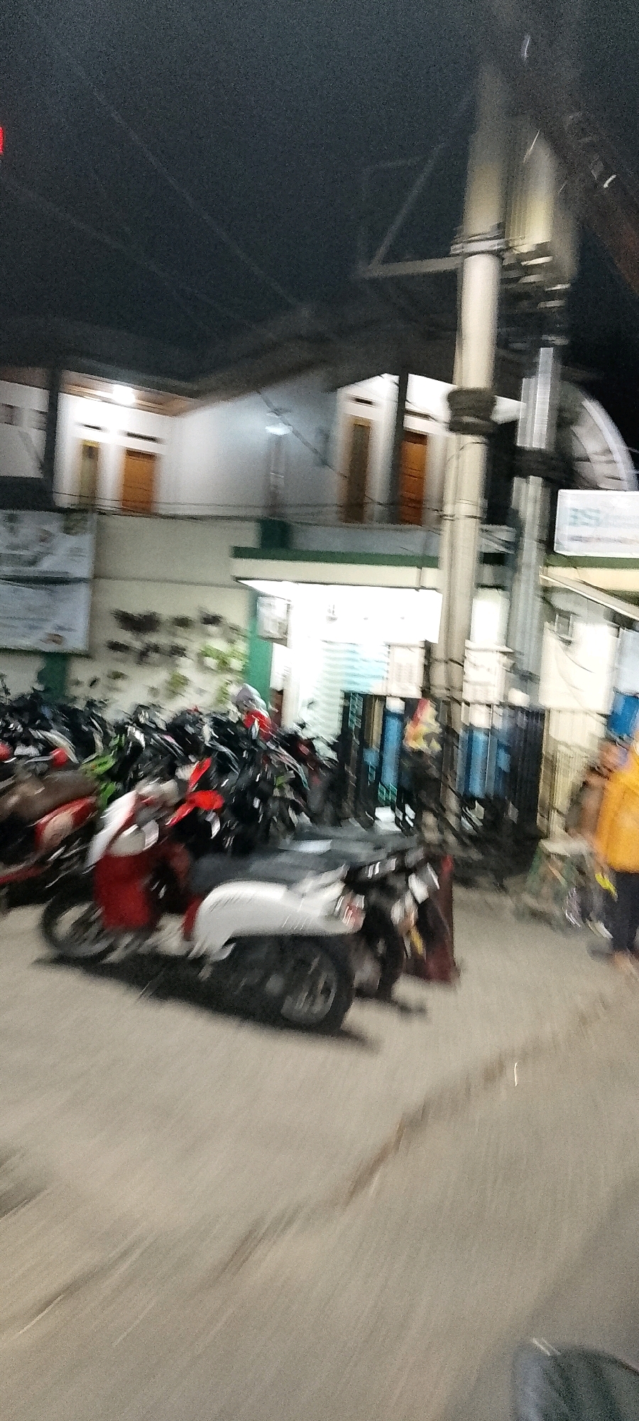 The image depicts a street scene in Karangsatria, Indonesia, at night, with several parked motorcycles. A multi-story building with white walls and brown doors is visible in the background, illuminated by interior lights. A tall pole, possibly a utility pole, stands prominently in the mid-ground, with some fencing behind it. In the foreground, various motorcycles are parked, with one prominent scooter in red and white colors. A person in a yellow jacket is partially visible on the right side of the image, and another person in dark clothing is also present. The ground appears to be paved. The image quality is low, with motion blur evident, suggesting movement during the capture.