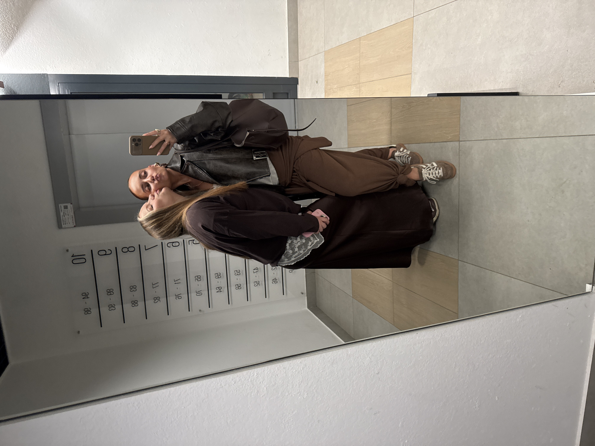 In a mirrored image, two women are positioned inside what appears to be the entryway of an apartment building, identified by the mailboxes aligned on the wall adjacent to them. The woman taking the photo with her smartphone is seen wearing a brown leather jacket and matching pants, and a pair of Adidas sneakers. The other woman has long hair, and she is wearing a brown dress with a lace overlay. The location of Izhevsk, Russia, is not confirmed.