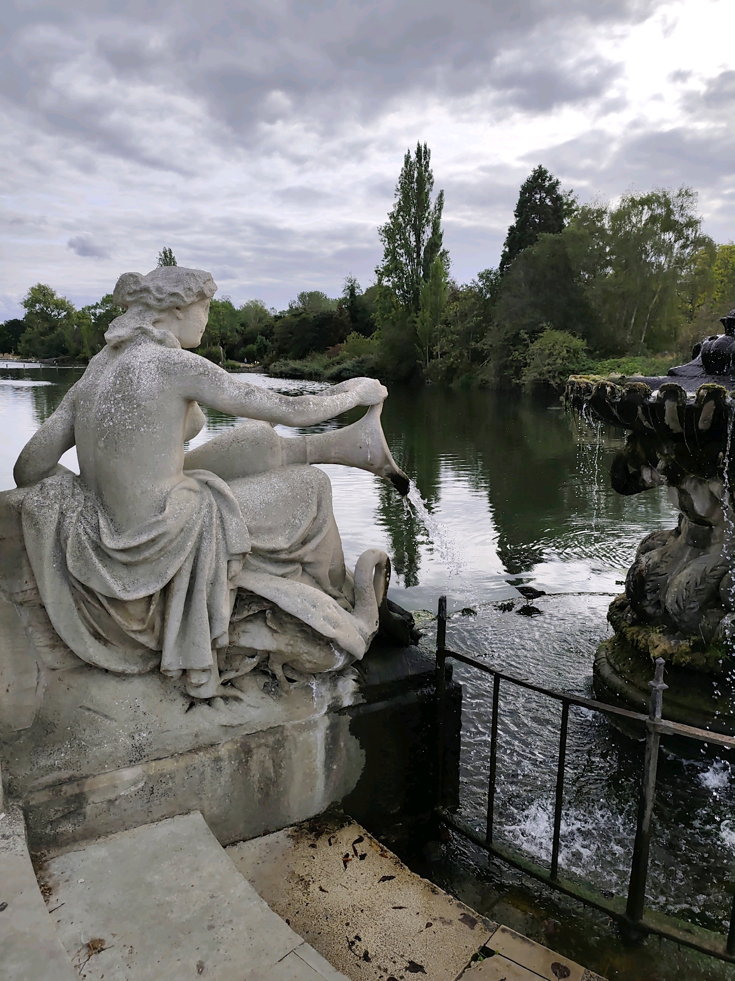 A stone statue of a reclining woman is situated next to a body of water in London. The statue is made of weathered stone and depicts the woman pouring water from a jug into the lake. She is seated on a plinth with a swan at her side. The water appears to be flowing from a fountain on the right side of the image, which is also made of stone and is partially covered in moss. The surrounding environment includes trees and a cloudy sky. The scene is tranquil and appears to be in a park or garden setting.