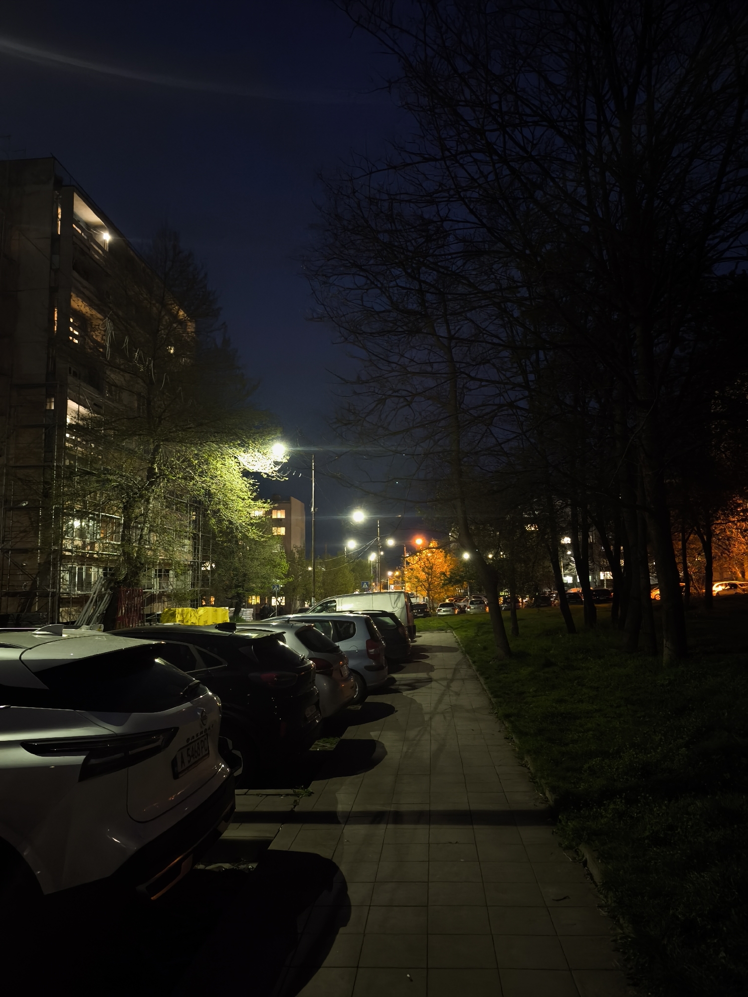 The image depicts a street scene at night in Burgas, Bulgaria. A multi-story building is visible on the left, partially covered in scaffolding. Cars are parked along the street, illuminated by streetlights. A sidewalk runs along the right side of the frame, bordered by a strip of grass and a line of trees. The sky is dark, and the overall lighting is low, creating long shadows. One car has a visible license plate, reading "A 5468 PL".