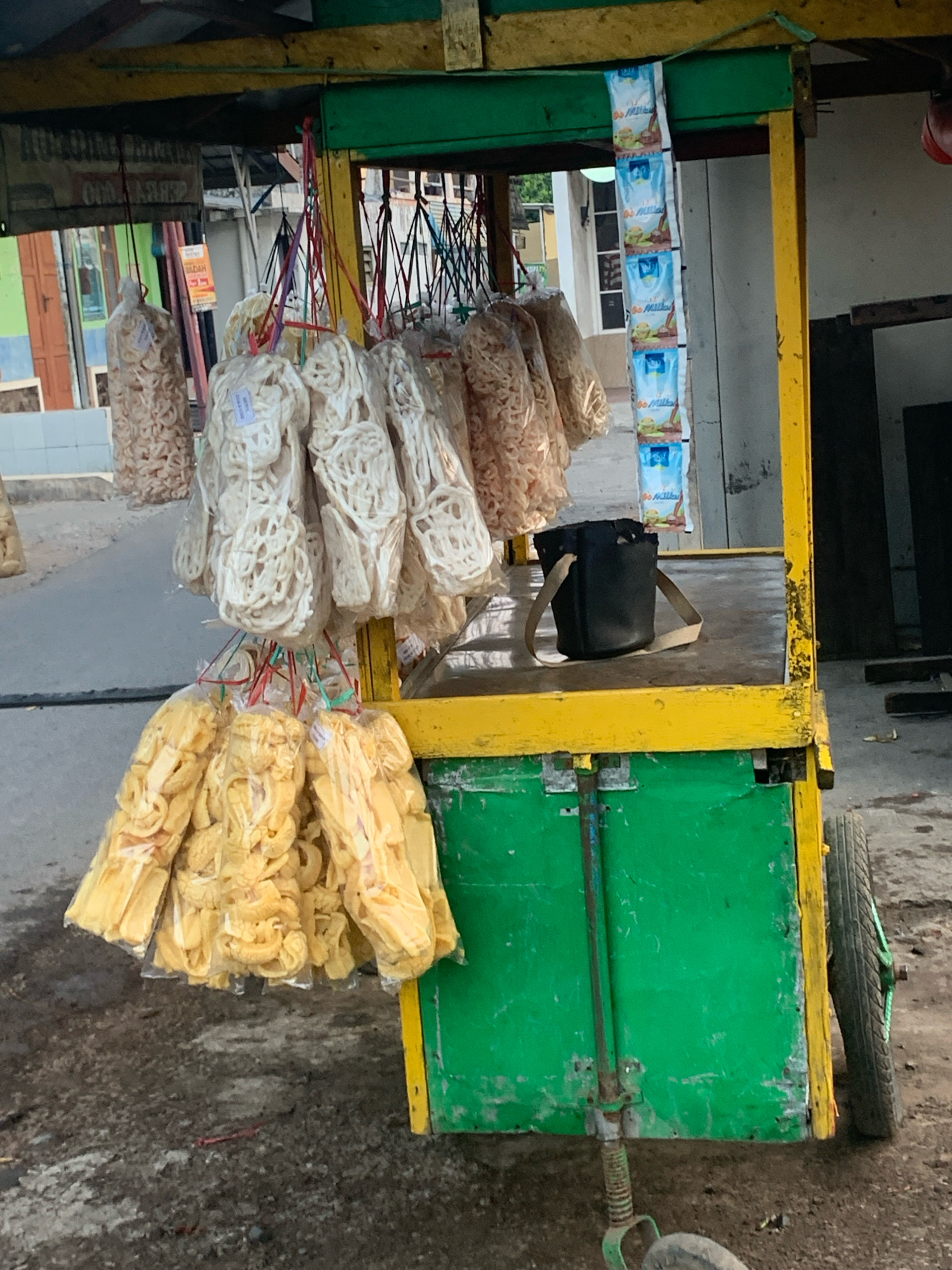 This is a medium-shot, eye-level photograph of a food cart selling snacks, likely located in Bululawang, Indonesia. The cart is painted bright green and yellow and has two wheels, one visible on the right. The cart's surface is laden with various packaged snacks, mostly light-colored and crispy, hanging in plastic bags. There is a black bag with a tan strap resting on the cart's surface. In the background, buildings with green and brown facades are visible, as well as a street with a dusty, worn surface. The lighting suggests it is daytime, possibly late afternoon. No people are visible in the frame.