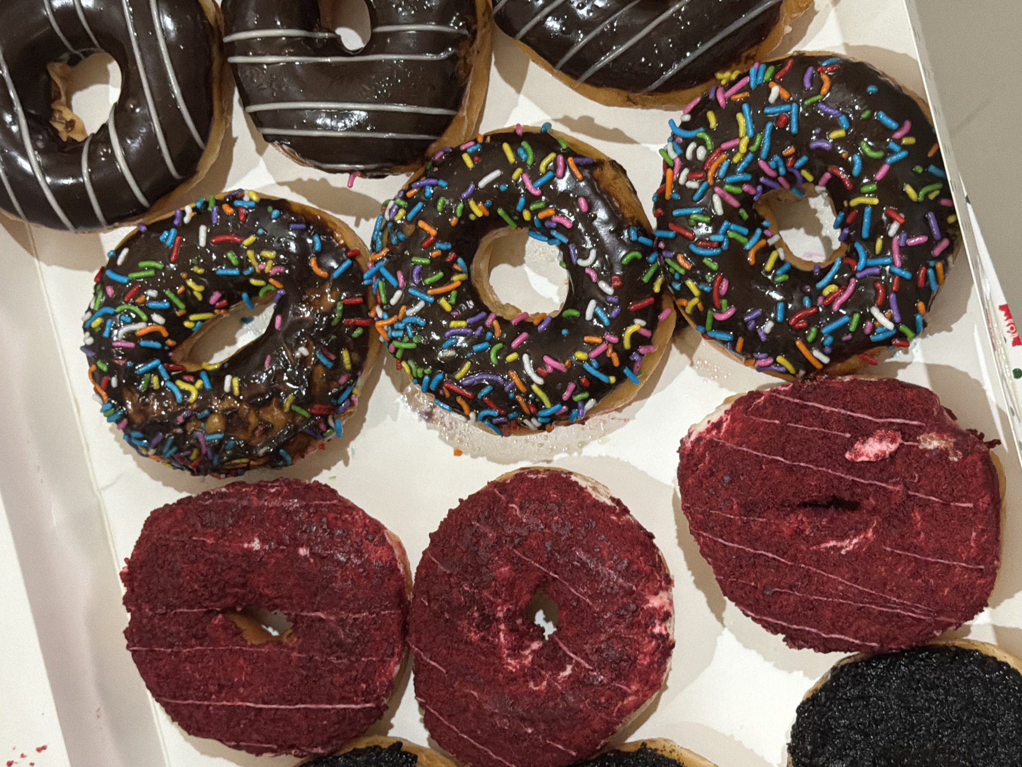 A top-down, high-angle shot depicts a box of donuts. The box contains three chocolate-frosted donuts with rainbow sprinkles, two chocolate-frosted donuts with white frosting lines, and three red velvet donuts. The donuts are arranged in a grid pattern within the white cardboard box. The photograph does not offer enough information to pinpoint the location in East Jakarta, Indonesia. There is no visible information about people, actions, interactions, or any specific environment.