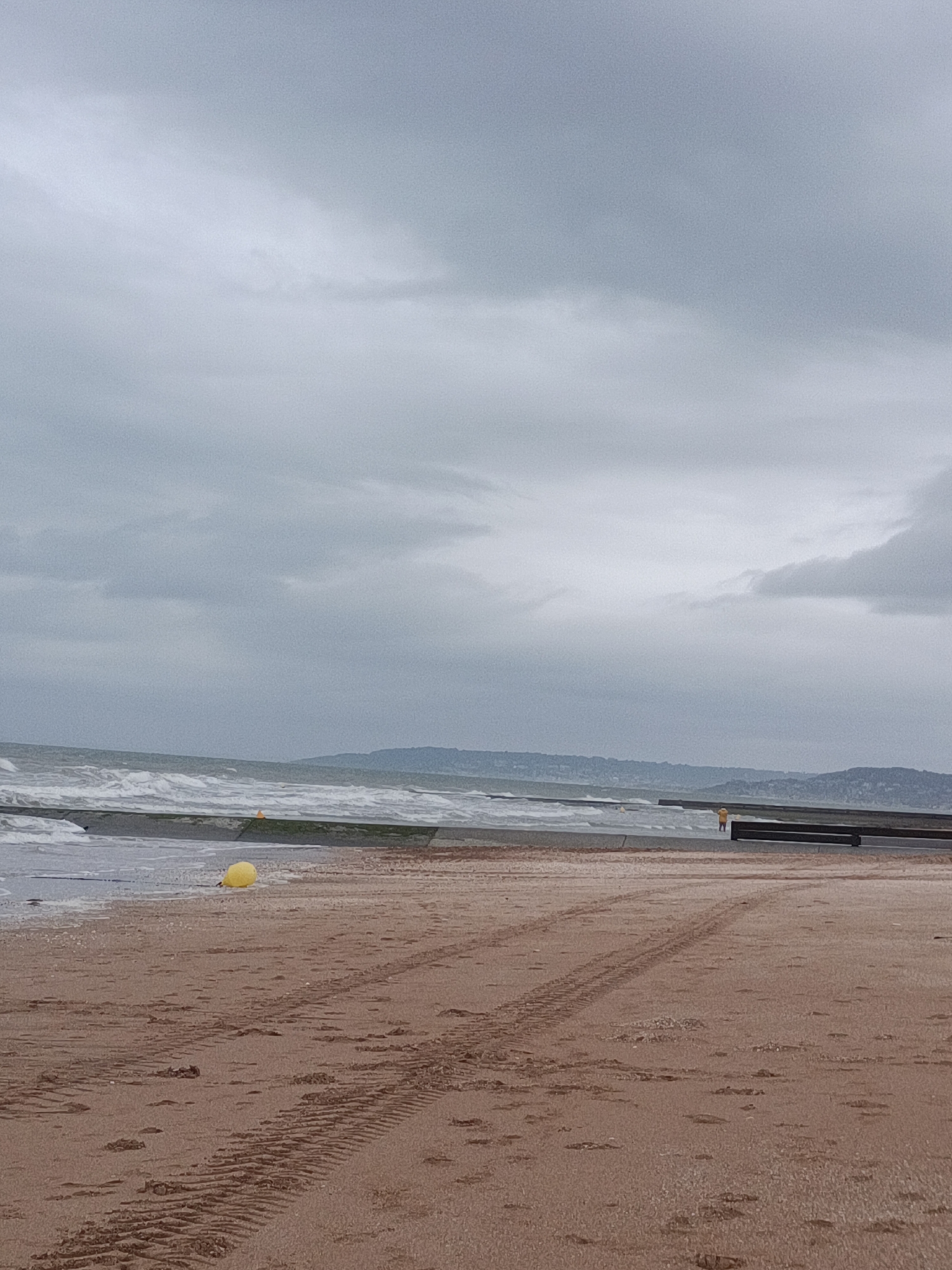 The image depicts a sandy beach in Houlgate, France, under a heavily overcast sky. The foreground is dominated by wet sand with tire tracks and footprints. A yellow buoy is visible on the sand near the waterline. In the middle ground, a concrete structure runs along the edge of the sea, with waves breaking against it. A person in a yellow garment stands on the beach near a wooden structure in the distance. The background features a hazy coastline with rolling hills. The overall scene is somber and muted due to the cloudy weather.