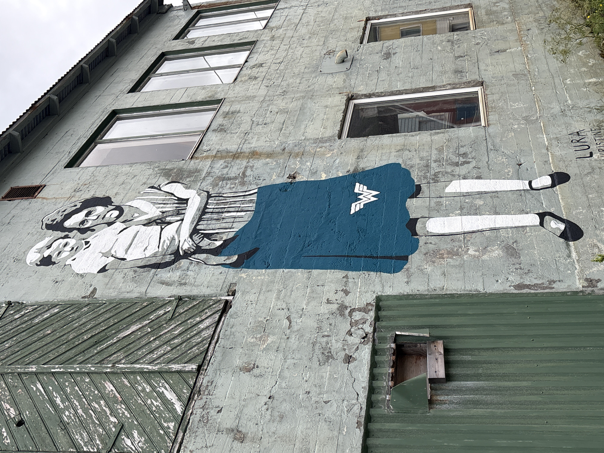 The image shows a mural painted on the exterior of a weathered concrete building in Gryllefjorden, Norway. The mural depicts a woman carrying a child, rendered in black and white with a vibrant blue skirt. The woman is dressed in a striped top and her hair is pulled back. The child is wearing white clothing. A stylized white "W" emblem, reminiscent of Wonder Woman's insignia, is visible on the blue skirt. The building itself has several windows and a corrugated green metal structure on the left side, which appears to be an extension or an attached shed. The concrete wall is textured and shows signs of wear, with patches of peeling paint and cracks. The roofline is visible at the top of the building. The overall scene is stark, with the mural providing a strong visual element against the utilitarian architecture. The mural is signed "LURA KRISTINE" at the bottom right.