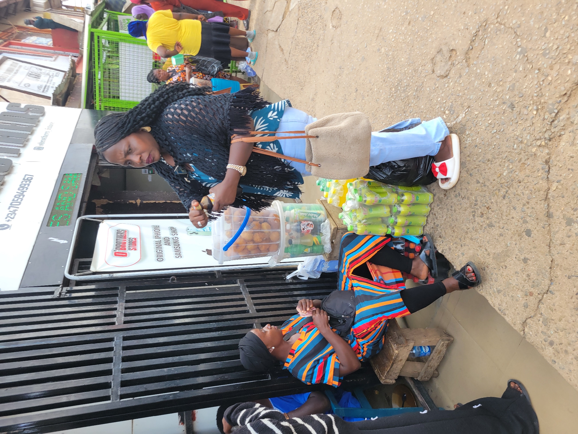 The image shows a street scene in Jos, Nigeria. A woman with braids and a black shawl stands near a display of drinks and food in plastic containers. She has a tan purse slung over her shoulder and wears blue jeans. Another woman sits on a small wooden stool nearby, wearing a colorful striped top and black leggings. She has a black head covering. Behind them, a shop sign reads "O'Brothers Stores" and advertises original iPhones and Samsung products. Other people are walking in the background. The pavement is visible, and there is a metal gate in the foreground.