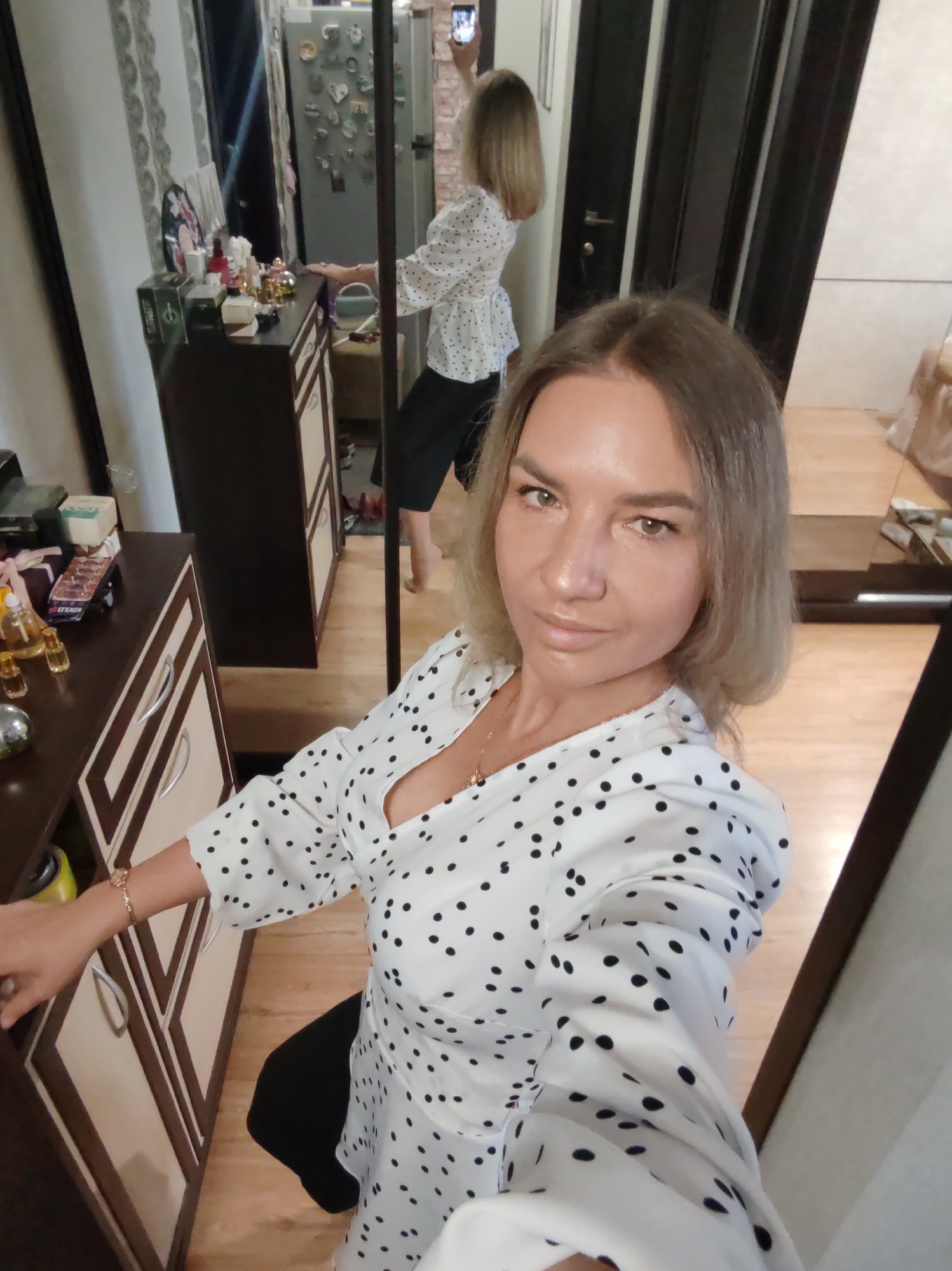 The image depicts a woman taking a selfie in an indoor setting, likely a home in Petropavlovsk-Kamchatsky, Russia. The woman, with shoulder-length light brown hair and a neutral to slightly pleasant expression, looks directly into the camera. She is wearing a white V-neck top with black polka dots, dark pants, a gold bracelet on her left wrist, and a gold necklace.
The photograph is taken in front of a large mirror, which provides a full reflection of the scene. In the reflection, the woman is seen holding a smartphone in her right hand to capture the image. Behind her in the reflection, there's a dark brown dressing table or cabinet with lighter wood accents and silver handles. The table is adorned with numerous items, including various small bottles (likely perfumes or cosmetics), a black box with green text that appears to read "ETRAEN" and a pink/purple packet possibly marked "BETAEH" (Cyrillic for BЕТАЕН).
Further in the background of the reflection, a light-colored refrigerator covered with many magnets of various shapes, including hearts, is visible. Adjacent to the refrigerator, a dark wooden door or doorway can be seen. The room features light wooden laminate flooring and a patterned wallpaper. Another dark brown cabinet is partially visible in the foreground to the left. The setting suggests a casual, domestic environment, likely during daytime hours.