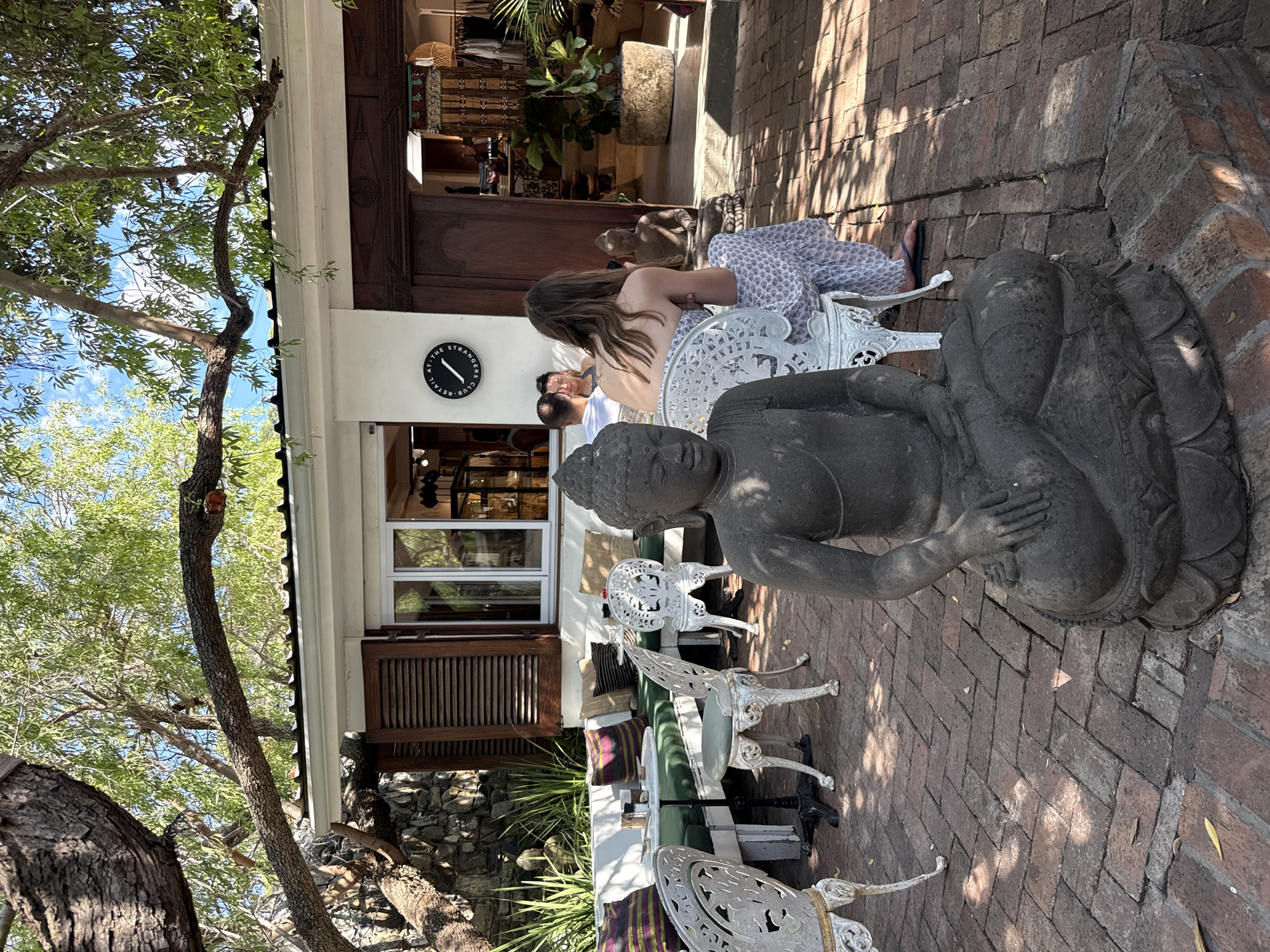 Here is a detailed description of the image:
The photograph captures a moment at "The Stranger's Club" retail store in Cape Town, South Africa. The scene unfolds in an outdoor patio area adjacent to the building. The patio is paved with brick, and a mature tree with extensive branches frames the left side of the shot.
Foreground:
A stone statue of Buddha sits prominently in the right foreground, casting a shadow on the brick patio.
Several white, ornate metal chairs are scattered around. Some chairs are placed near tables, suggesting a café or seating area.
Midground:
People are seated at one of the tables. Two women are visible, one with long hair. There is a man sitting at the table, wearing a white shirt.
A building is in the background. The white-walled portion has a clock and a window. The building is otherwise dark wood.
Background:
The store front is visible in the background, showcasing clothing and other merchandise. Green plants add a touch of nature.
Overall, the setting evokes a relaxed and stylish atmosphere.