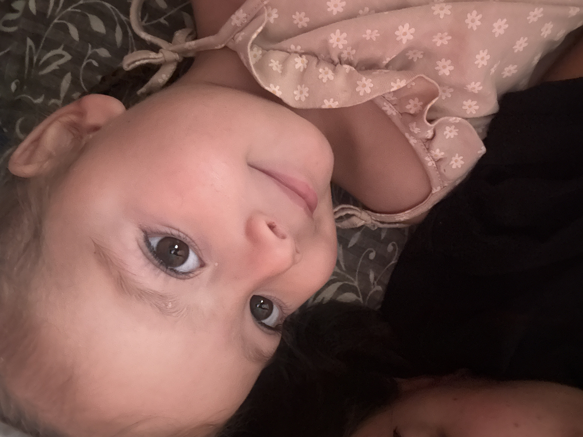A close-up, overhead, portrait-style photograph shows a young child lying on their side, looking directly at the camera. The child has fair skin, light brown hair, and large, dark brown eyes. They are wearing a light pink dress adorned with small white flowers. The background is a dark, textured surface, possibly a blanket or bedding. The child's head is resting on a dark surface, likely an adult's shoulder or arm, with a portion of another person's face visible in the lower left corner of the image. The image appears to be taken indoors in a dimly lit environment. There are no visible landmarks or elements in the image that can be used to determine the location in Basarabeasca, Moldova.