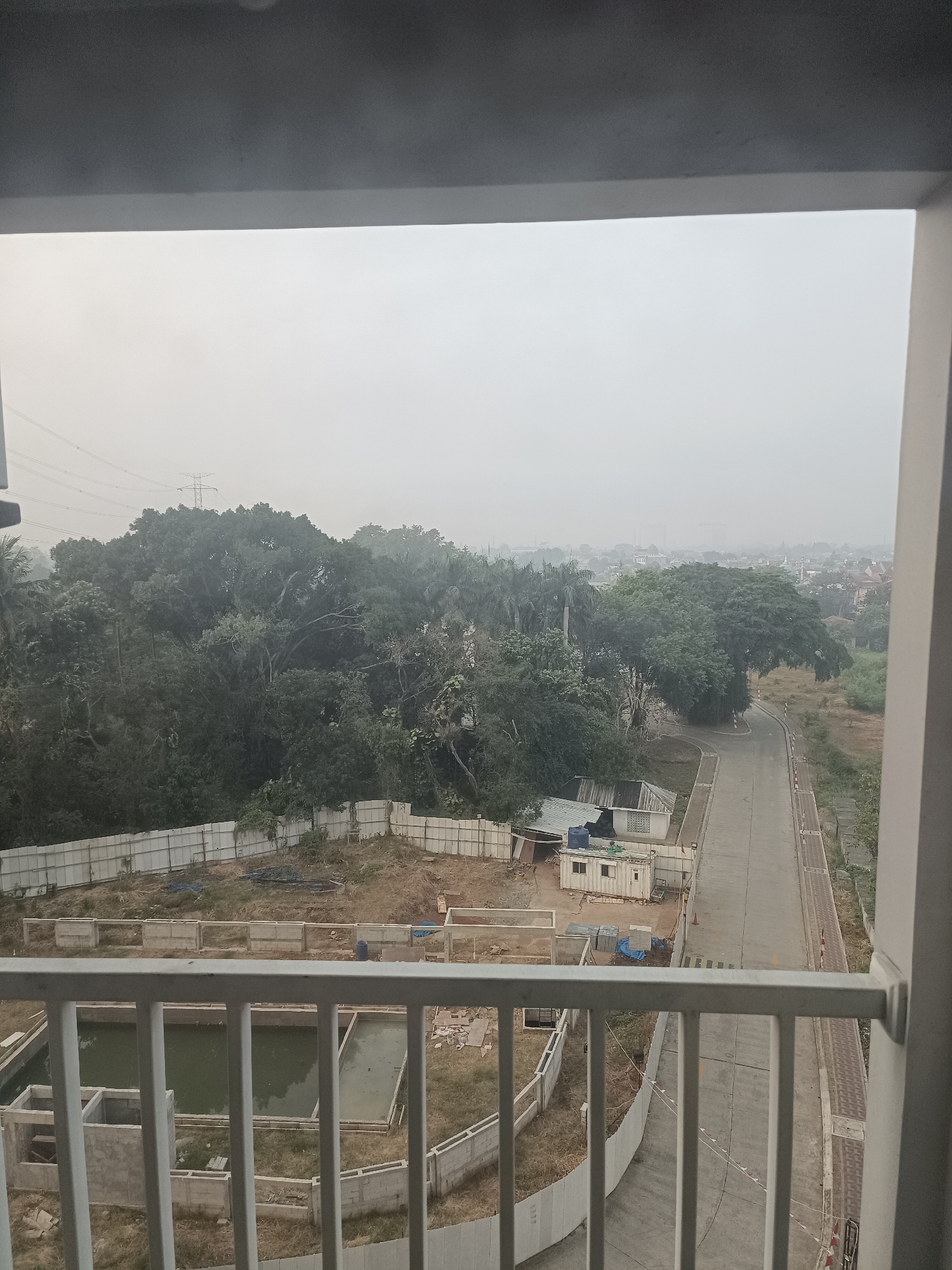 The image depicts a view from a balcony overlooking a developing area in South Tangerang, Indonesia. In the foreground, a white metal railing is visible. Beyond the railing, there are two rectangular water tanks under construction, with concrete structures and murky green water.
The midground shows a construction site with a white fence enclosing an undeveloped plot of land. Several portable buildings, including a white container-like structure with a blue water tank next to it, are present on the site. A paved road curves through the scene, with some construction materials and barriers along its edges.
The background is dominated by a dense forest of lush green trees, with a power transmission tower and lines visible in the hazy sky above. Further in the distance, a cityscape with buildings can be seen through the atmospheric haze, suggesting a semi-urban or peri-urban environment. The overall impression is one of ongoing development amidst a natural landscape, under an overcast or hazy sky.