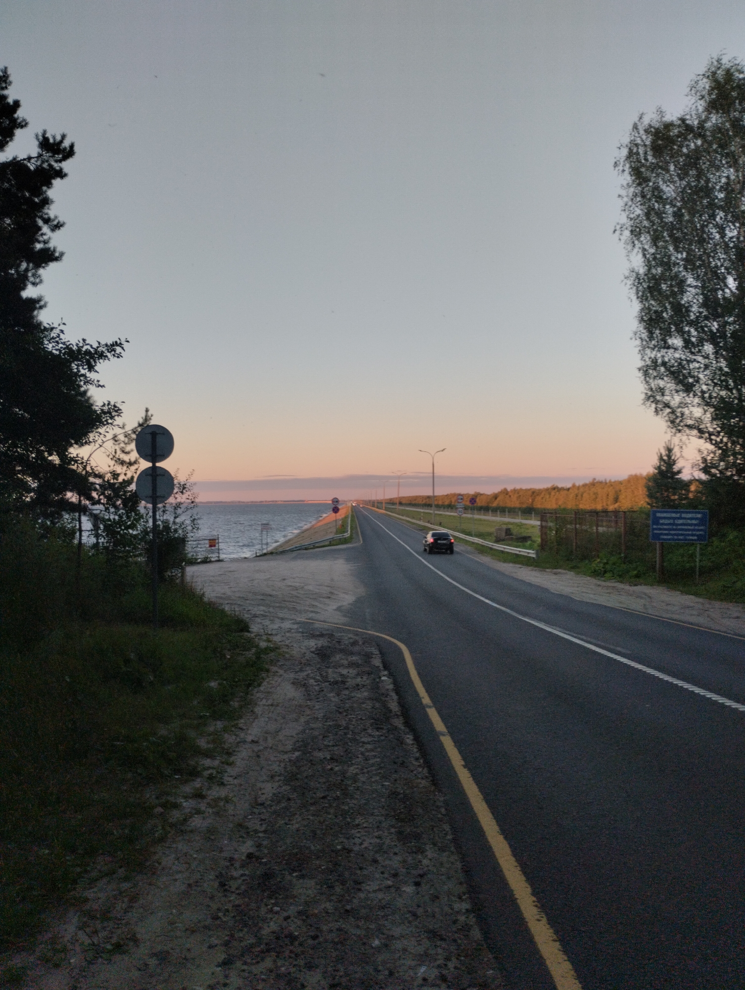 The image captures a scenic view of a highway running alongside a body of water, likely Lake Gorky, on the outskirts of Zavolzhye, Russia, during what appears to be sunset. The sky is a gradient of pale blue and soft orange, indicating the time of day.
On the left side of the frame, the dark silhouettes of trees dominate the foreground, with their branches reaching towards the sky. Adjacent to the trees is a dirt and gravel path, leading to the water's edge. Two circular traffic signs on a pole are visible near the water.
The highway, paved with asphalt, stretches into the distance, marked by white lines. A single black car is driving on the right lane. The left side of the highway is bordered by a fence and a line of trees, with a blue informational signpost visible in the distance on the right side of the road. The water is calm, reflecting the soft hues of the sky. The overall atmosphere is serene and quiet, typical of a rural highway at dusk.