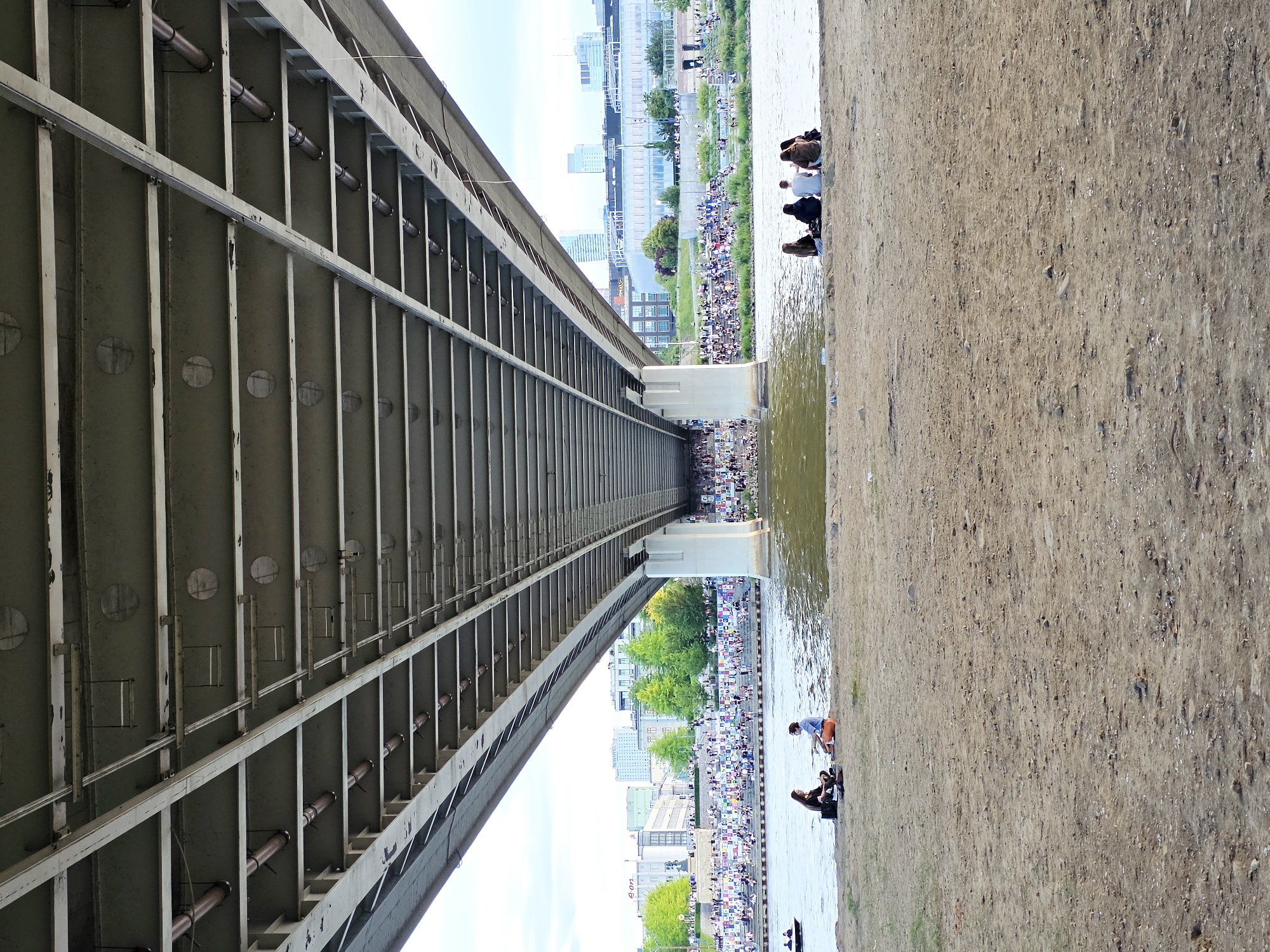 Underneath the vast expanse of a bridge, a sandy riverbank hosts several groups of people relaxing by the water. In the foreground, two individuals sit facing each other on the sandy shore, engaged in conversation. Further along the bank, a cluster of five people are seated in a semi-circle, facing the river. The river itself is calm, with a small boat visible in the distance on the left. On the opposite bank, a bustling scene unfolds. A large crowd is gathered on tiered seating or grassy slopes, overlooking the river. Trees dot the landscape, and a city skyline with modern buildings forms the backdrop. The underside of the bridge is a complex structure of metal beams and pipes, painted in muted tones. The scene suggests a leisurely gathering in an urban setting, likely a popular spot for relaxation and people-watching by the Vistula River in Warsaw, Poland.