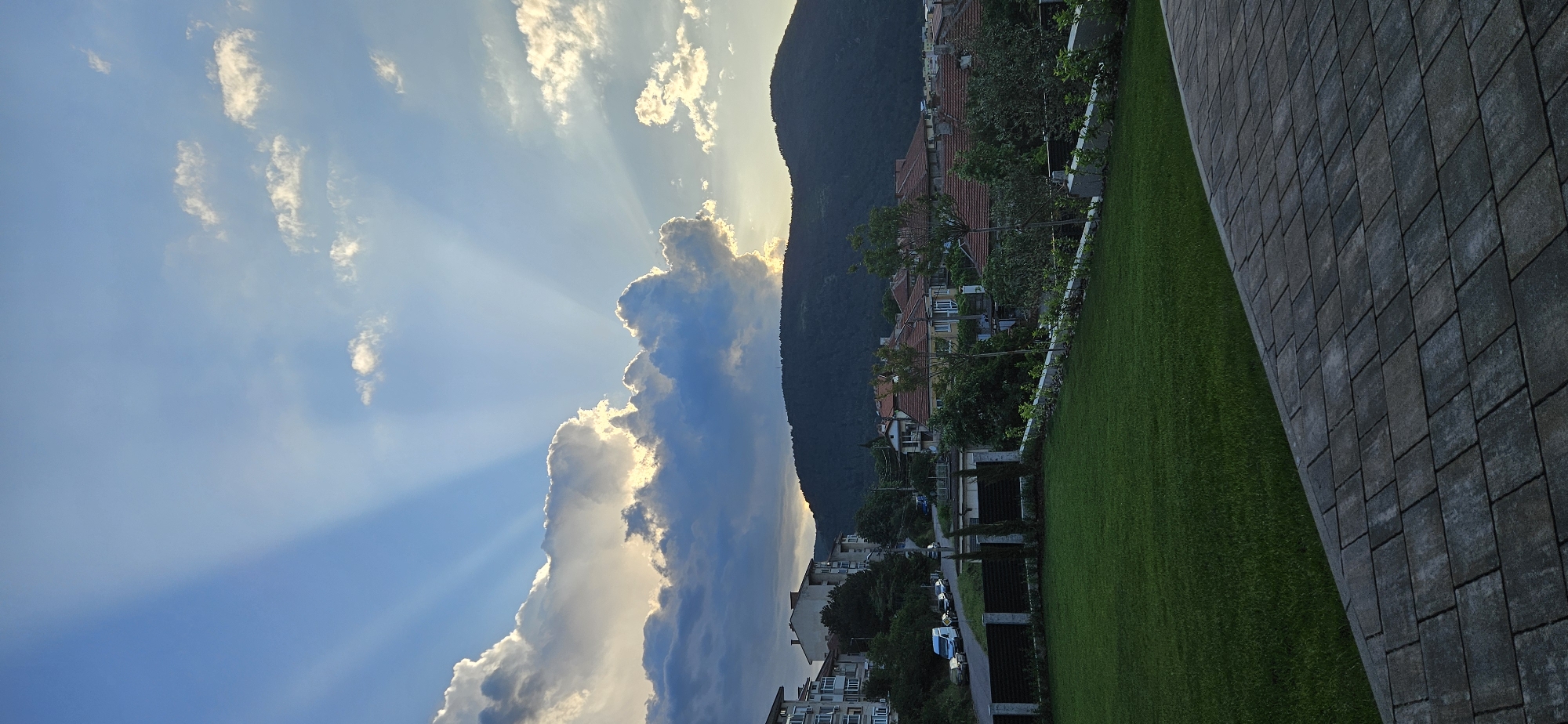 The image presents a view of a townscape under a partially cloudy sky, marked by crepuscular rays shining through the clouds. A dense, verdant mountain rises adjacent to a cluster of buildings, their red-tiled roofs indicative of residential structures. A green lawn stretches from the base of these buildings, meeting a paved area constructed with gray bricks. The positioning of the photo suggests a view from an elevated position.