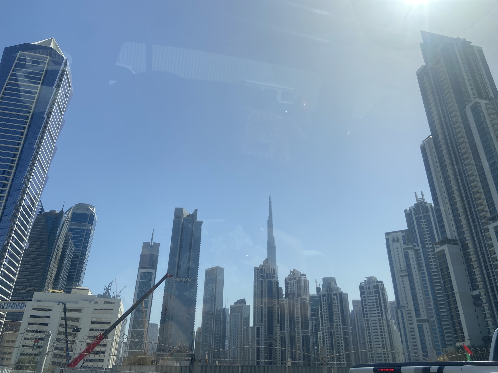The image captures a daytime view of Dubai's Business Bay skyline, United Arab Emirates, as seen from a vehicle's interior. Several high-rise buildings dominate the scene, including the Burj Khalifa, which is centrally positioned within the cluster of skyscrapers. The buildings exhibit diverse architectural styles. A construction crane is visible amidst the urban landscape. Fencing is visible in the foreground. The sky is clear and bright, with the sun's glare creating a bright spot at the top of the image. The interior of the vehicle creates a reflective effect.