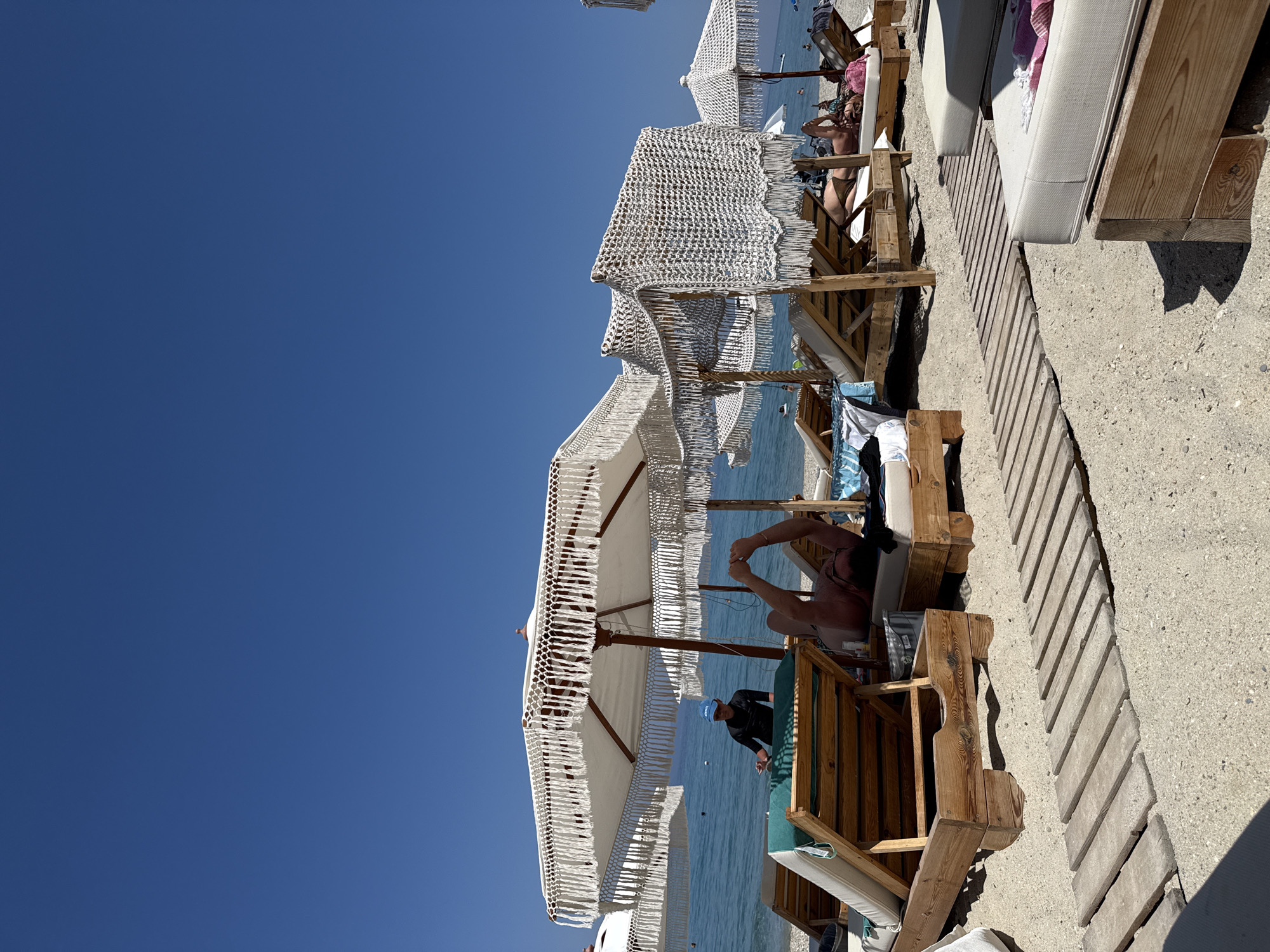 The image depicts a sunny day at a beach resort in Pallini Municipal Unit, Greece. The scene is oriented vertically, with the deep blue sky occupying the left side of the frame. In the foreground and middle ground, several wooden lounge chairs are set up on a sandy and pebbly beach, shaded by white umbrellas with decorative fringe. The chairs are occupied by people relaxing. One person is lying on a lounge chair under an umbrella, with their arms raised. Another person, wearing a blue cap and a dark top, is standing near the water.
The beach overlooks a calm, blue sea. The overall atmosphere is one of leisure and relaxation, indicative of a vacation setting. The bright sunlight suggests it is midday or afternoon. The presence of lounge chairs and umbrellas points to a well-established beach resort or a similar tourist area. No visible text is present in the image.