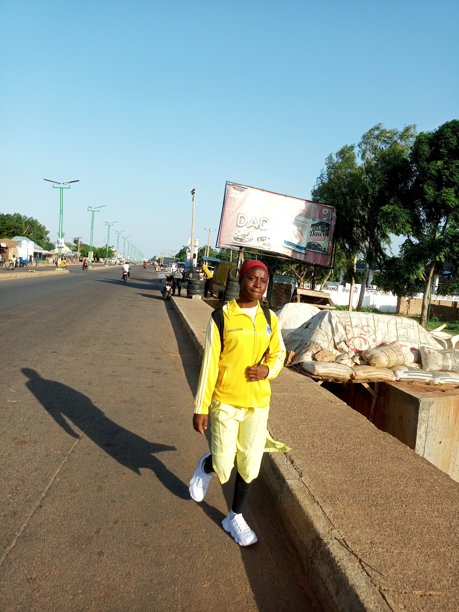 A young woman wearing a yellow tracksuit and white sneakers stands on the side of a road in Garko, Nigeria. She is looking towards the camera. The road is wide and appears to be a main thoroughfare with multiple lanes of traffic and streetlights. In the background, there are shops, buildings, trees, and a billboard. Several people and vehicles, including motorcycles and cars, are visible on the road and sidewalks. Piles of what appear to be sandbags or cement sacks are stacked on the right side of the road. The sky is clear and blue, suggesting it is daytime. The overall scene depicts a busy urban environment in Nigeria.