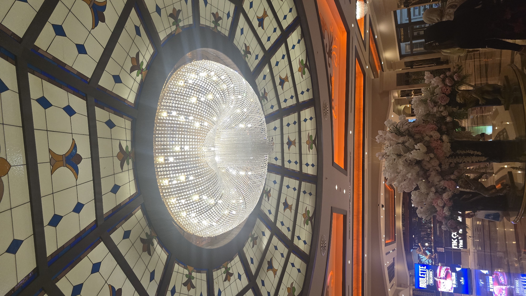 The image shows the interior of a building, likely a casino or hotel lobby, in Kazafani, Northern Cyprus. A large, ornate crystal chandelier hangs from a stained-glass ceiling. The ceiling features a geometric pattern with blue diamonds and floral motifs in yellow and green. Below the ceiling, there are brightly lit orange architectural elements. In the foreground, a large floral arrangement is displayed on a circular table with reflective surfaces. To the left, a digital screen advertises a "ROULETTE TOURNAMENT." In the background, there are entrances to various establishments, including one labeled "LUXURY BRAND." Several individuals are visible in the background, including two women facing away from the camera. The overall impression is one of opulence and high-end entertainment.