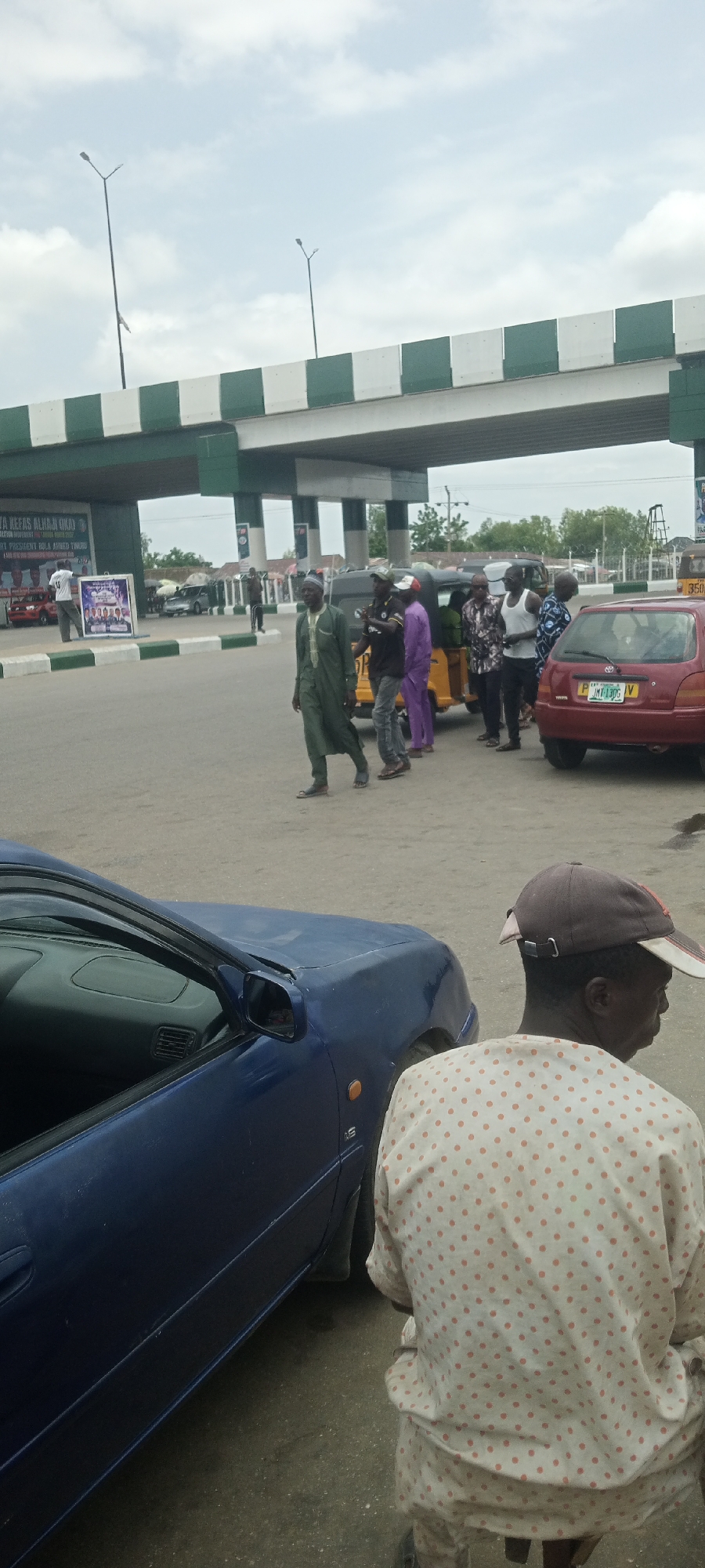 The image depicts a street scene in Jalingo, Nigeria, under an overcast sky. In the foreground, a person wearing a light-colored, polka-dotted shirt and a cap is partially visible, facing away from the camera. To the left, the front portion of a dark blue car is seen. Behind this car, a group of people are gathered near a yellow auto-rickshaw and a red car. Some of the individuals are dressed in traditional African attire, including a man in a long green garment. A man in a purple garment is also present. In the background, a multi-level structure, likely an overpass or bridge, with green and white patterned barriers runs across the scene. A large banner with text and images is visible on the left side of the structure, indicating a local event or political campaign. Streetlights are also visible above the overpass. The overall environment suggests a public area with moderate activity.