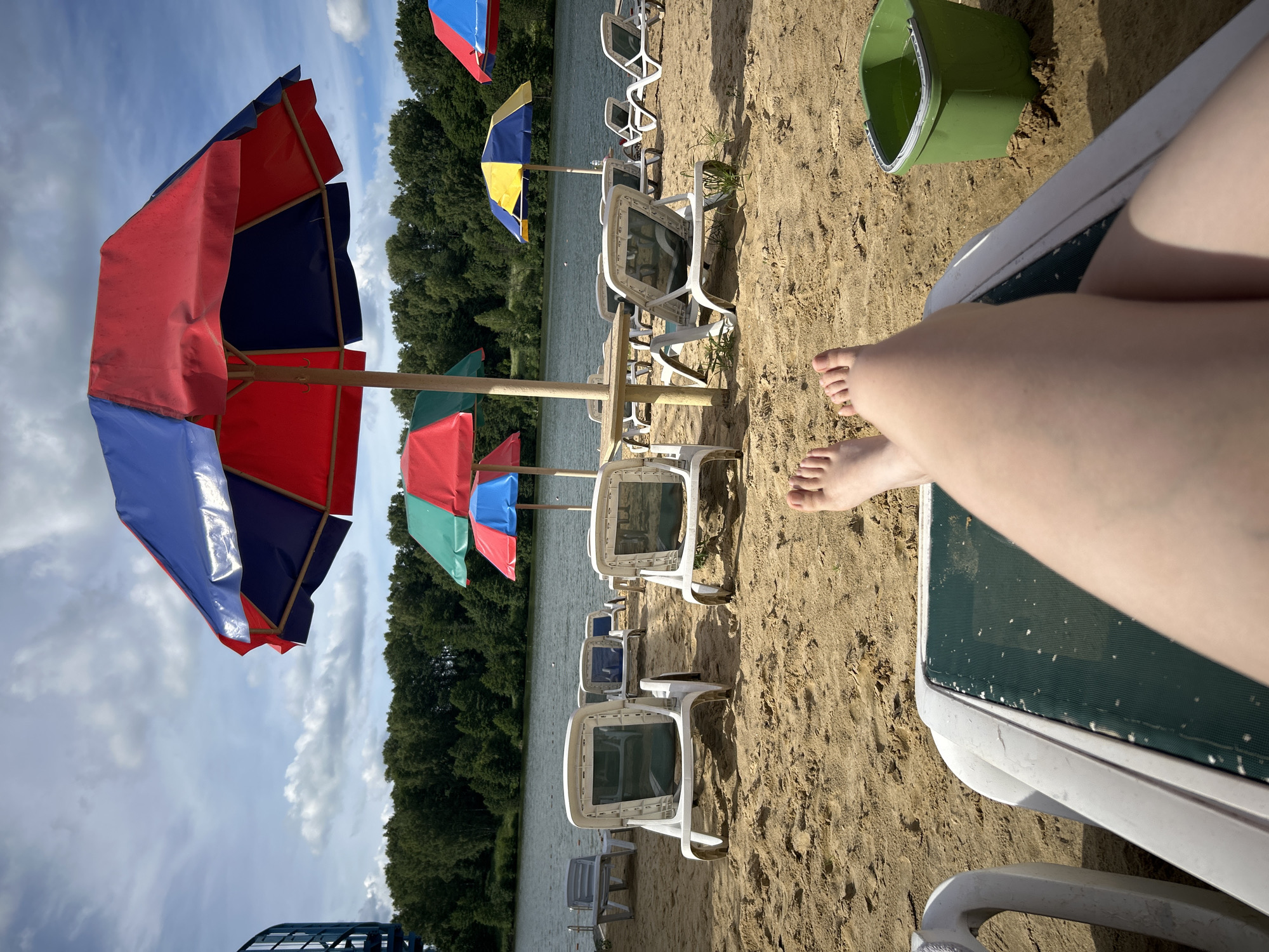 The image depicts a sun-drenched sandy beach adjacent to a calm body of water, likely a lake, bordered by a dense line of trees. Multiple rows of white plastic lounge chairs and colorful beach umbrellas are arranged on the sand, suggesting a public recreational area. In the foreground, a pair of bare feet are visible, resting on a green and white striped lounge chair. To the right of the chair, a bright green plastic bucket is partially filled with water and sand, placed on the beach. The sky is partly cloudy, with diffused sunlight casting shadows on the sand and illuminating the scene. The overall impression is one of a tranquil summer day at a beach resort, potentially located in or near the city of Yubileynyy, Russia, given the geographical context provided in the prompt.