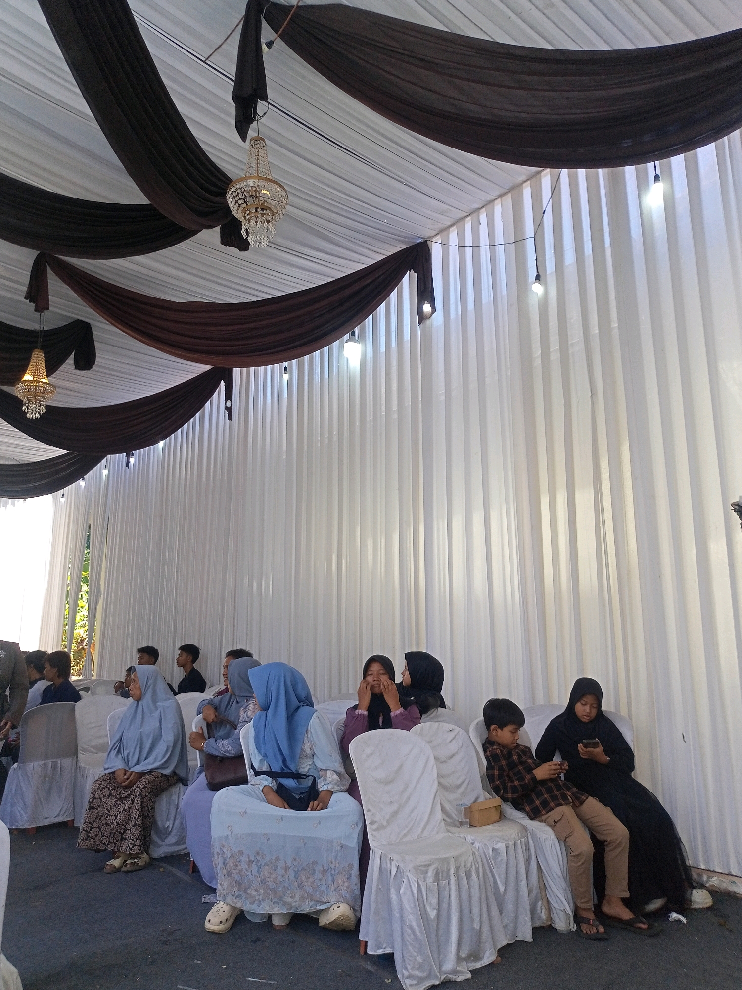 A wedding reception is taking place outdoors under a tent in Limbangan, Indonesia. The tent's ceiling is draped with brown and white fabric, with two ornate chandeliers hanging from the top. The sides of the tent are lined with white curtains. Several people are seated on white chairs facing the camera, some appearing to be guests. The floor is covered with a dark carpet.