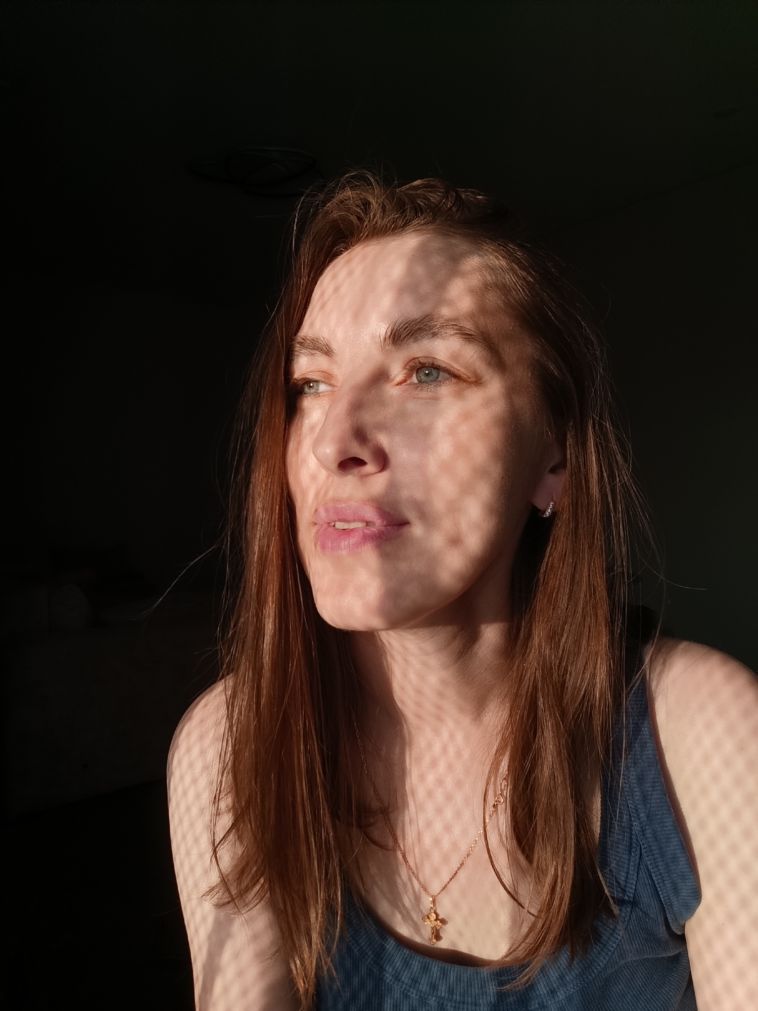 A medium shot shows a woman with light brown hair looking to the left. The woman is wearing a blue tank top and a gold necklace with a cross pendant. The lighting in the image is dramatic, with harsh shadows cast across the woman's face and body, suggesting she is near a window with blinds or a lattice structure. The background is dark and out of focus. The image was taken in Chaikovsky, Russia.