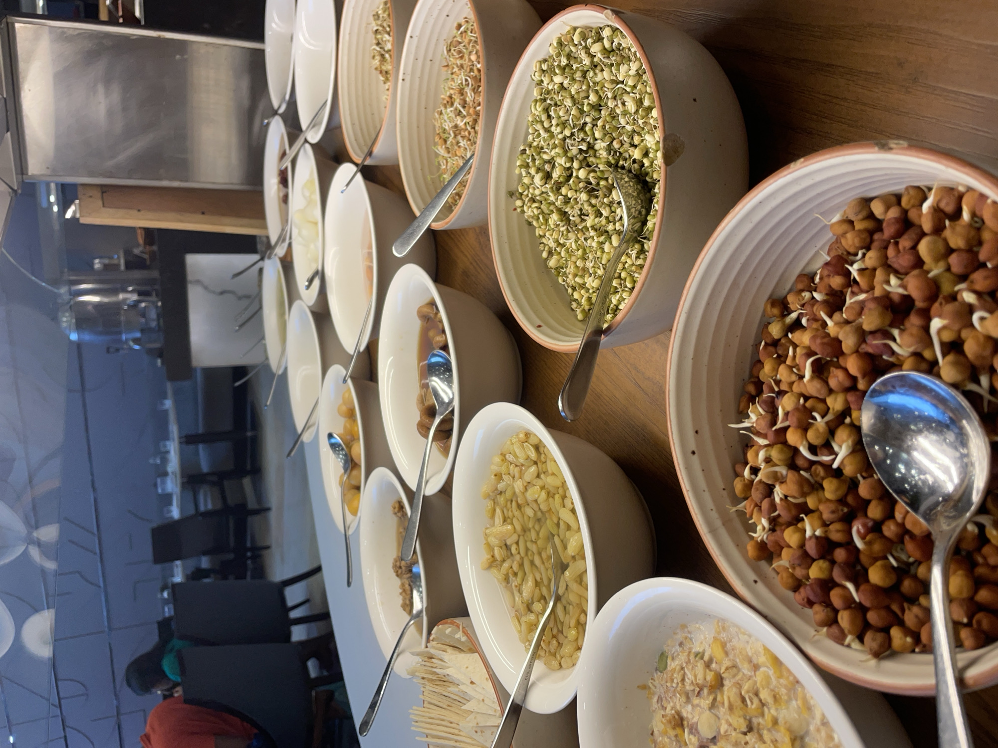 In a well-lit indoor setting, possibly a restaurant or hotel buffet, a variety of food items are displayed in white ceramic bowls on a wooden table. The scene is dominated by bowls containing sprouted legumes and grains, such as green moong sprouts, sprouted chickpeas in various shades of brown and red, and other unidentified sprouted seeds. Some bowls contain what appear to be pickled or stewed fruits or vegetables, with a dish of what looks like tortilla chips or flatbread on the side. Spoons are present in several bowls, indicating that the food is meant to be served. In the background, blurred figures of people are visible, along with tables and chairs, suggesting a dining area. The overall impression is one of a breakfast or light meal buffet. There is no indication of the city of Agra, India in the image.