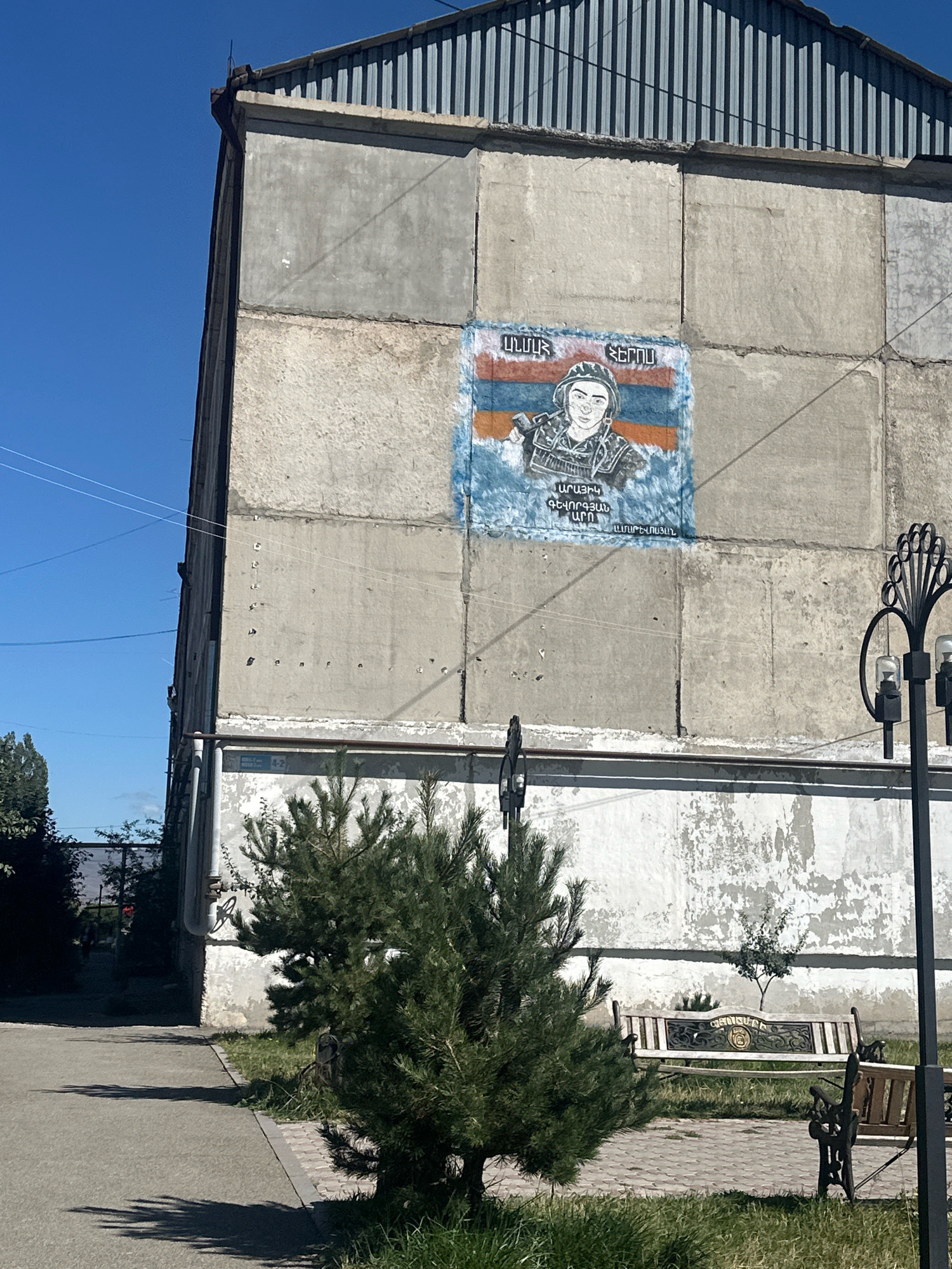 The image captures a mural on the side of a concrete building, likely located in Gyumri, Armenia, given the Armenian inscriptions on the mural. The mural depicts a person in military attire, holding a rifle, with the Armenian flag prominently displayed behind them. The building itself appears to be old, with weathered concrete walls and a corrugated metal roof. In the foreground, a paved walkway runs alongside a grassy area with a small pine tree. A decorative black lamppost stands to the right of the mural. There are no people actively present in the scene, suggesting a public space or the exterior of a building. The sky is clear and blue, indicating a daytime setting. The overall scene conveys a sense of remembrance or tribute.