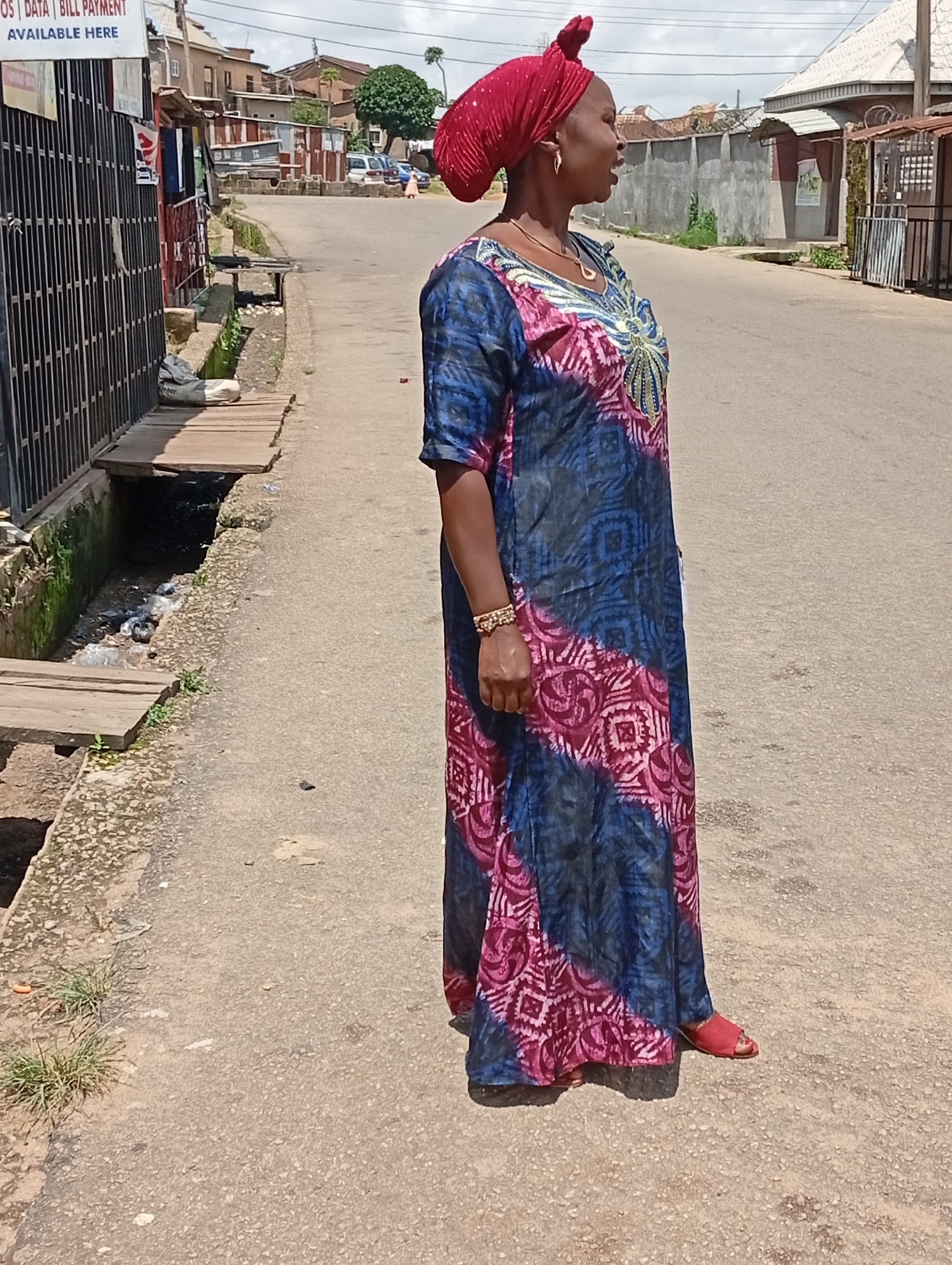 The image depicts a woman standing on a paved road in Jos, Nigeria. She is wearing a red headscarf and a long, colorful dress with a blue and pink tie-dye pattern. She has a gold necklace and a bracelet on her right wrist. The woman is facing to the left of the frame, with her body angled slightly towards the camera.
To the left of the road, there is a drainage ditch filled with debris, covered in part by wooden planks. Behind the ditch, a black metal fence encloses an area with various structures, including buildings with corrugated iron roofs and what appears to be a kiosk. A sign with text, including "AVAILABLE HERE", is visible on the fence. In the background, more buildings and a tree are visible, suggesting an urban or semi-urban environment. The road is asphalt and shows signs of wear. The sky is cloudy. The overall scene suggests a common street view in a Nigerian city.