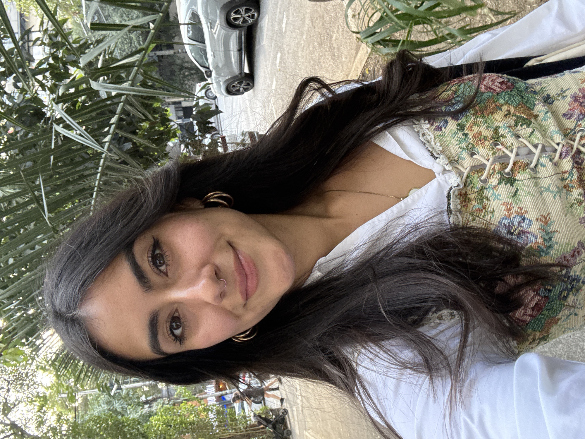 A young woman with dark hair and a nose ring is smiling slightly at the camera. She is wearing gold hoop earrings, a white shirt, and a tapestry-style corset top with lace-up detailing. The background features green foliage, a sidewalk, and a blurred view of a street with cars. The location is likely in Mexico City, Mexico, based on the plants and architecture visible in the background.