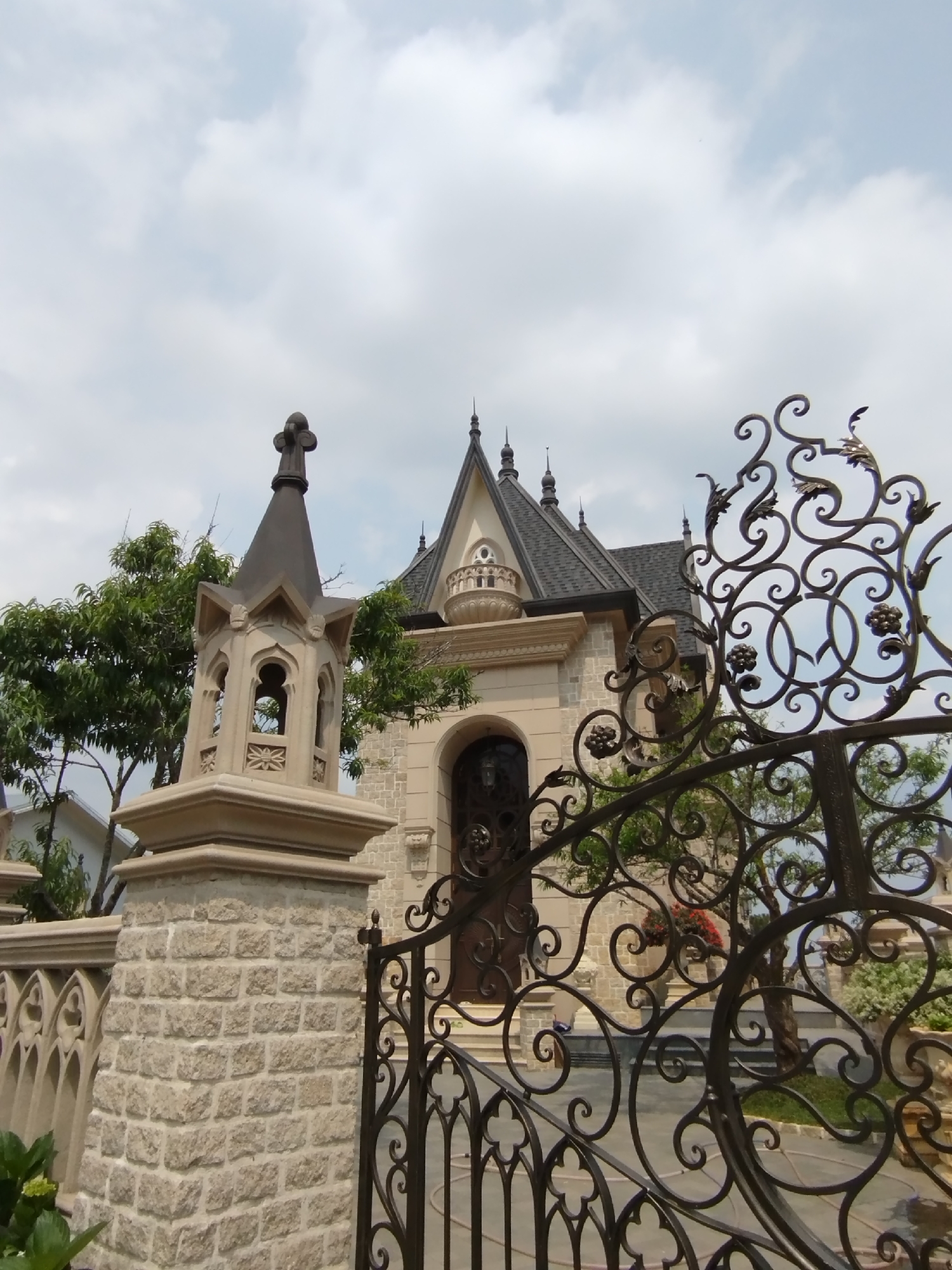 This image presents a grand, European-style house or villa, likely in Da Lat, Vietnam, viewed through an ornate wrought iron gate. The foreground is dominated by this dark metal gate, intricately designed with elaborate scrolls, leafy motifs, and clusters resembling grapes, along with gothic-inspired quatrefoil patterns along its lower edge.
To the left, a sturdy pillar made of light-colored, rough-hewn stone blocks supports a decorative stone structure. This architectural ornament resembles a miniature bell tower or finial, featuring arched openings and a dark, pointed roof topped with a cross or ornate finial.
Beyond the gate, the house is constructed from light-toned stone or stucco, exuding a classic aesthetic. It boasts a steeply pitched, multi-gabled roof covered in dark tiles, punctuated by several sharp spires and finials. A distinctive feature is a small, rounded turret or bay window on the upper left side of the house, complete with an arched window. The main entrance appears to have a dark, arched wooden door.
Green trees and bushes surround the property, suggesting a landscaped garden or natural setting. The sky is overcast with light clouds, indicating a daytime scene. The overall impression is one of timeless elegance and architectural sophistication, with no people or specific activities depicted. No text is visible in the image.