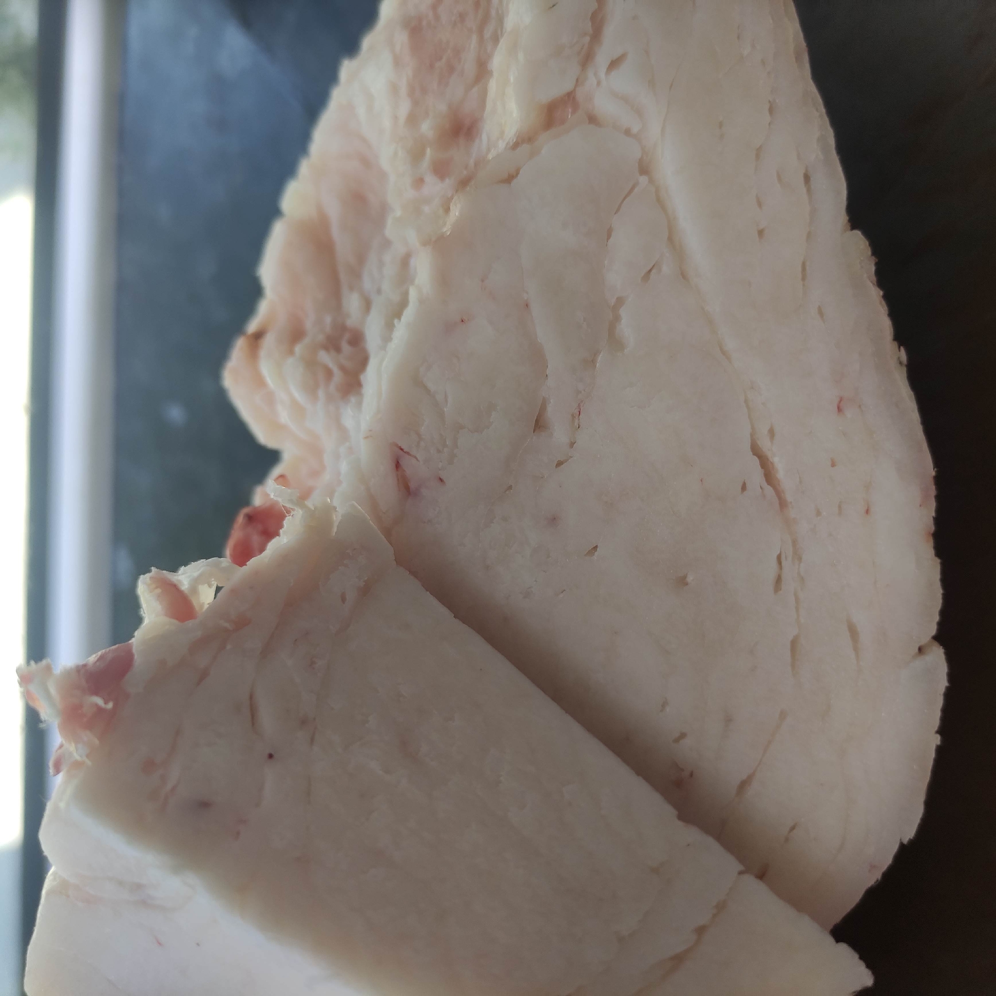 A close-up, medium shot captures a substantial piece of raw pork fat, likely lard, on a dark, textured surface. The fat is predominantly white with a creamy, opaque appearance, showing layers and a slightly waxy texture. Small, reddish streaks, possibly indicating blood vessels or remnants of meat, are visible throughout the fat. The surface it rests on is out of focus but appears to be a dark, smooth material, possibly a countertop or cutting board. The background is blurred and consists of a light horizontal band above a darker area, suggesting a window or a light source behind the subject, and a darker horizontal surface below. There are no people present, and no discernable actions are taking place beyond the static presentation of the food item. The lighting is soft and diffused, highlighting the texture and color of the fat. The image does not contain any information that would allow for its localization to Razzakov, Kyrgyzstan.