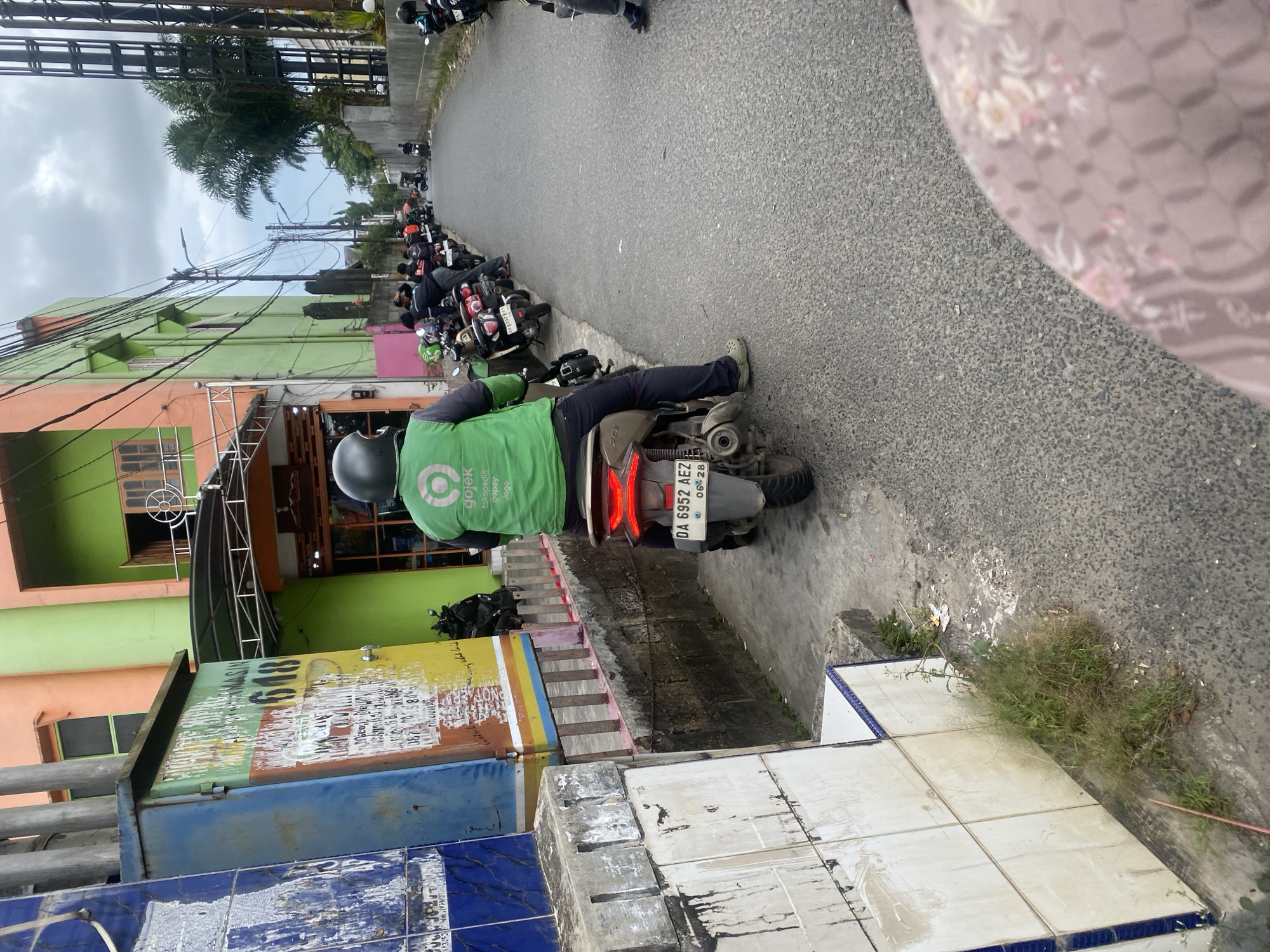 The image depicts a street scene in Sungai Paring, Indonesia. In the foreground, a person wearing a green Gojek uniform is seated on a scooter, facing away from the camera. The scooter has a license plate that reads "DA 6952 AEZ". The person is wearing a helmet. To the left of the scooter, there is a blue and yellow electrical box with faded markings and advertisements. Behind the electrical box, a building with orange and green facades is visible. Along the right side of the street, a row of parked scooters is lined up. The street is paved and appears to be a two-way road. In the background, several palm trees and a cloudy sky can be seen. The overall environment suggests a typical urban street in Indonesia, possibly a waiting area or a hub for ride-sharing services like Gojek.