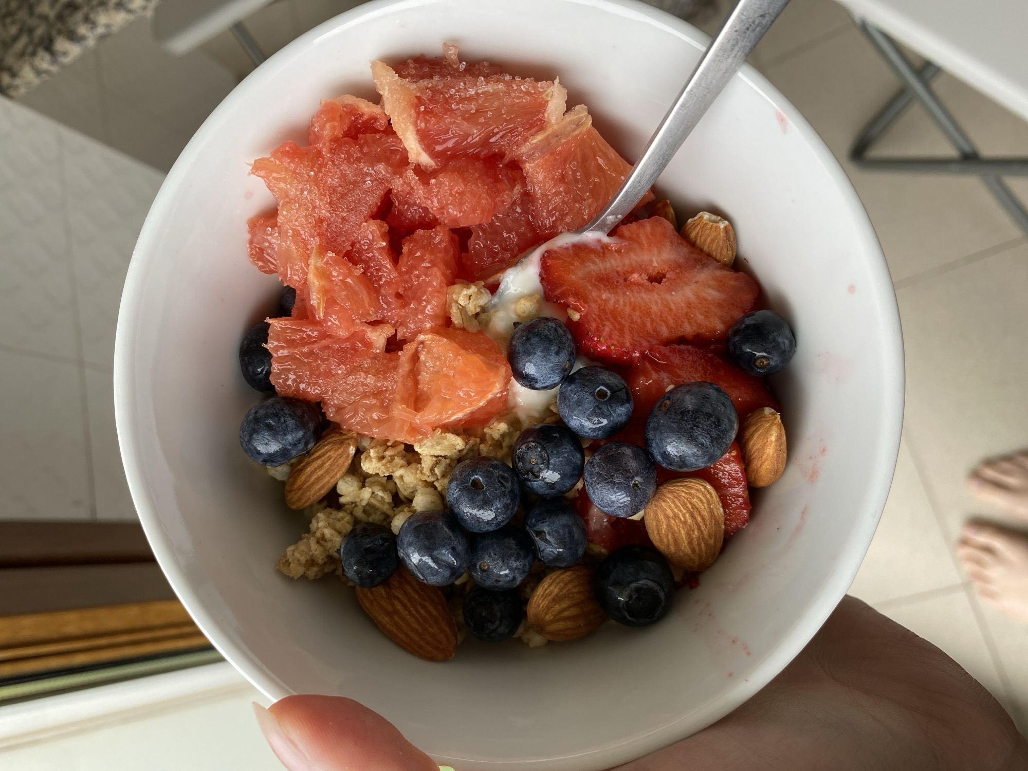 A close-up, high-angle shot depicts a white bowl held in a person's hand, filled with food. The bowl contains grapefruit segments, sliced strawberries, blueberries, almonds, and granola. A silver spoon is partially visible in the bowl. The background includes a kitchen counter and parts of chairs. There is no indication that the scene is in Gijón/Xixón, Spain.