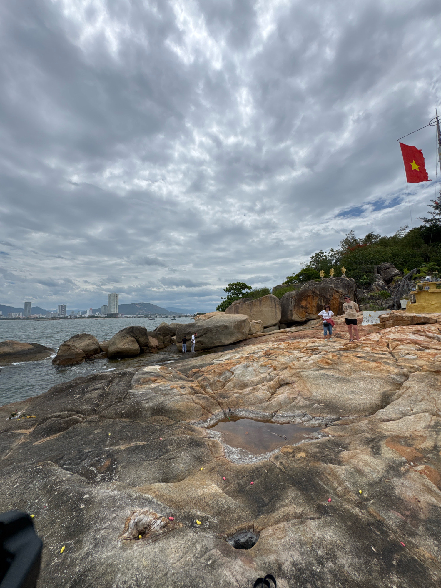 The image captures a rocky coastline in Nha Trang, Vietnam, under a cloudy sky. In the foreground, large, weathered rocks dominate the scene, with some showing evidence of small pools of water. The sea laps against the rocks on the left side of the frame.
In the mid-ground, a few people are visible. Two individuals, one wearing a tan t-shirt and shorts, and another in a white top and jeans, stand on the rocks. Further back, near the water's edge, two more figures can be seen.
The background reveals the Nha Trang cityscape with several tall buildings and mountains in the distance. A red flag with a yellow star, the flag of Vietnam, flies from a pole on a hill on the right side of the image. The hill is covered in lush greenery and features some structures, including golden statues on a large rock formation. The overall impression is one of a natural coastal landscape juxtaposed with urban development and cultural elements.