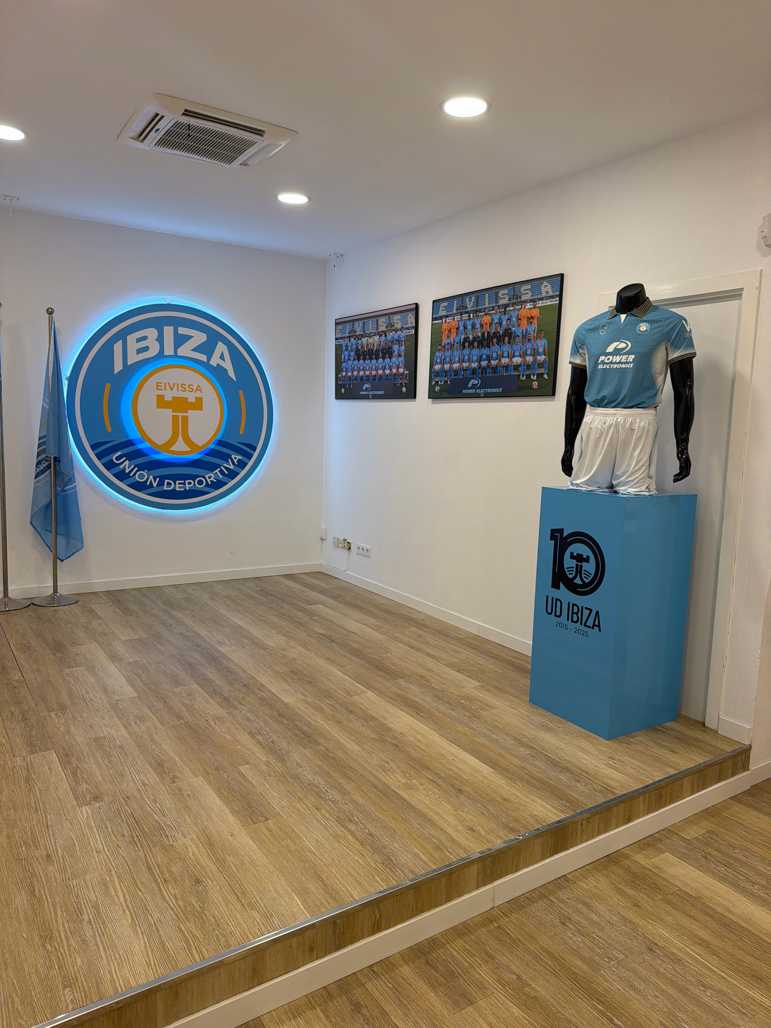 The image depicts an interior space, likely an office or a fan shop, within the city of Ibiza, Spain, exhibiting elements related to the UD Ibiza football club. Dominating the left wall is a large, illuminated circular logo of the club, featuring the text "IBIZA," "EIVISSA," and "UNION DEPORTIVA." To the left of the logo, a blue flag with a white logo is displayed on a silver flagpole.
On the right side of the room, two framed team photographs are mounted on the wall. Below these photos, a light blue football jersey and white shorts are displayed on a black mannequin, which stands on a light blue pedestal. The pedestal features a large "10" logo and the text "UD IBIZA 2015-2025," signifying the club's decade anniversary.
The room has light-colored walls and a wooden plank floor. A silver metal transition strip separates the floor at the entrance from the main area. An air conditioning unit is visible on the ceiling. The overall impression is one of a well-maintained and decorated space dedicated to the football club.