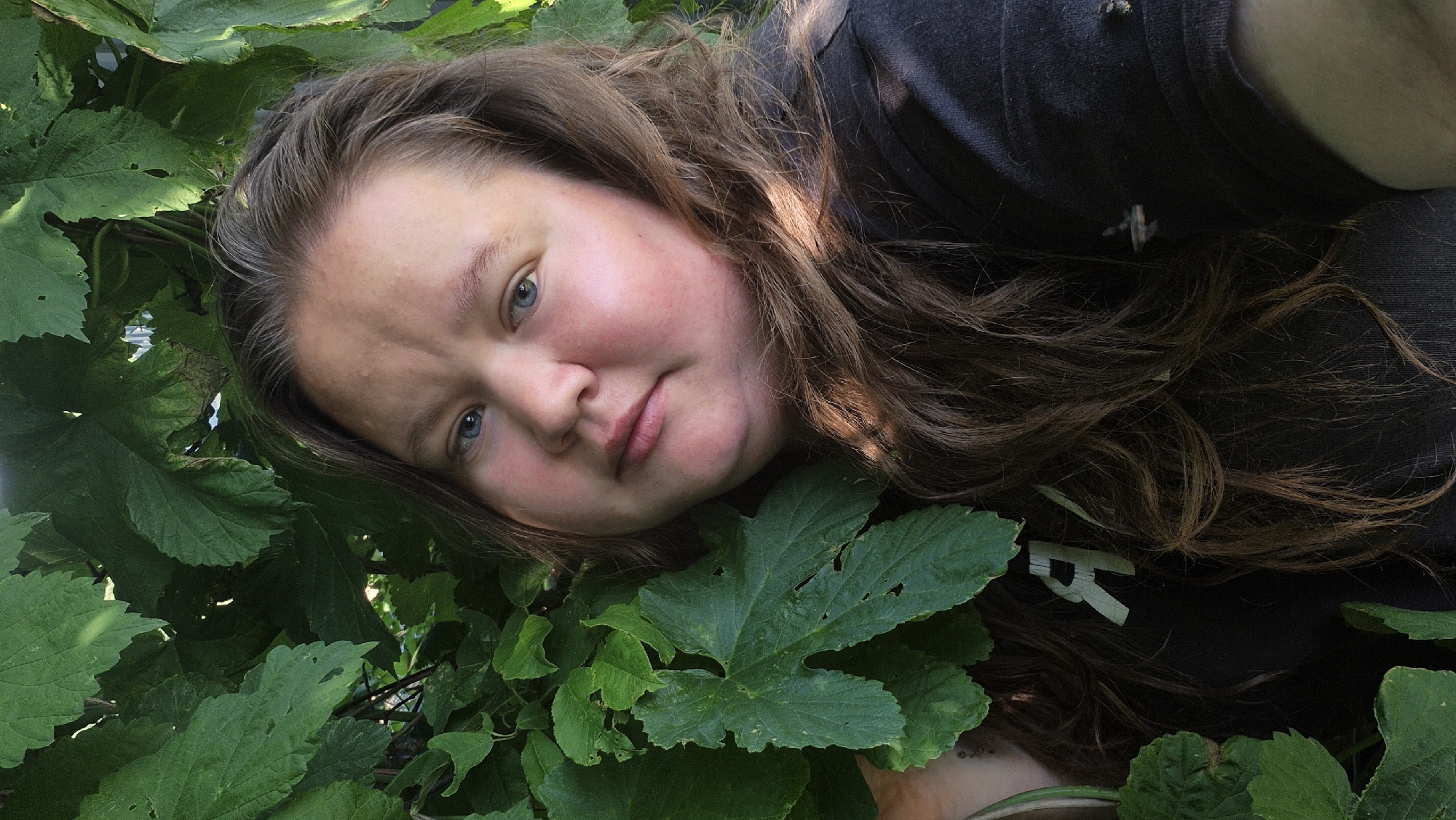 The media file shows a close-up, high-angle shot of a young person lying amongst lush green foliage, likely in a garden or natural outdoor setting. The individual has long, wavy brown hair and blue eyes, and appears to be looking directly at the camera with a relaxed and contented expression, with rosy cheeks suggesting warmth. They are wearing a dark t-shirt. The dense green leaves create a natural frame around the person's face and upper body, highlighting their features and the vibrant greenery. Sunlight filters through the leaves, casting soft shadows and illuminating parts of their face and hair, suggesting it is daytime. A faint logo or text, appearing to be "R", is visible on the dark fabric of the person's shirt. The overall impression is one of peacefulness and connection with nature, possibly taken in a rural setting like Дойная, Russia, given the context.