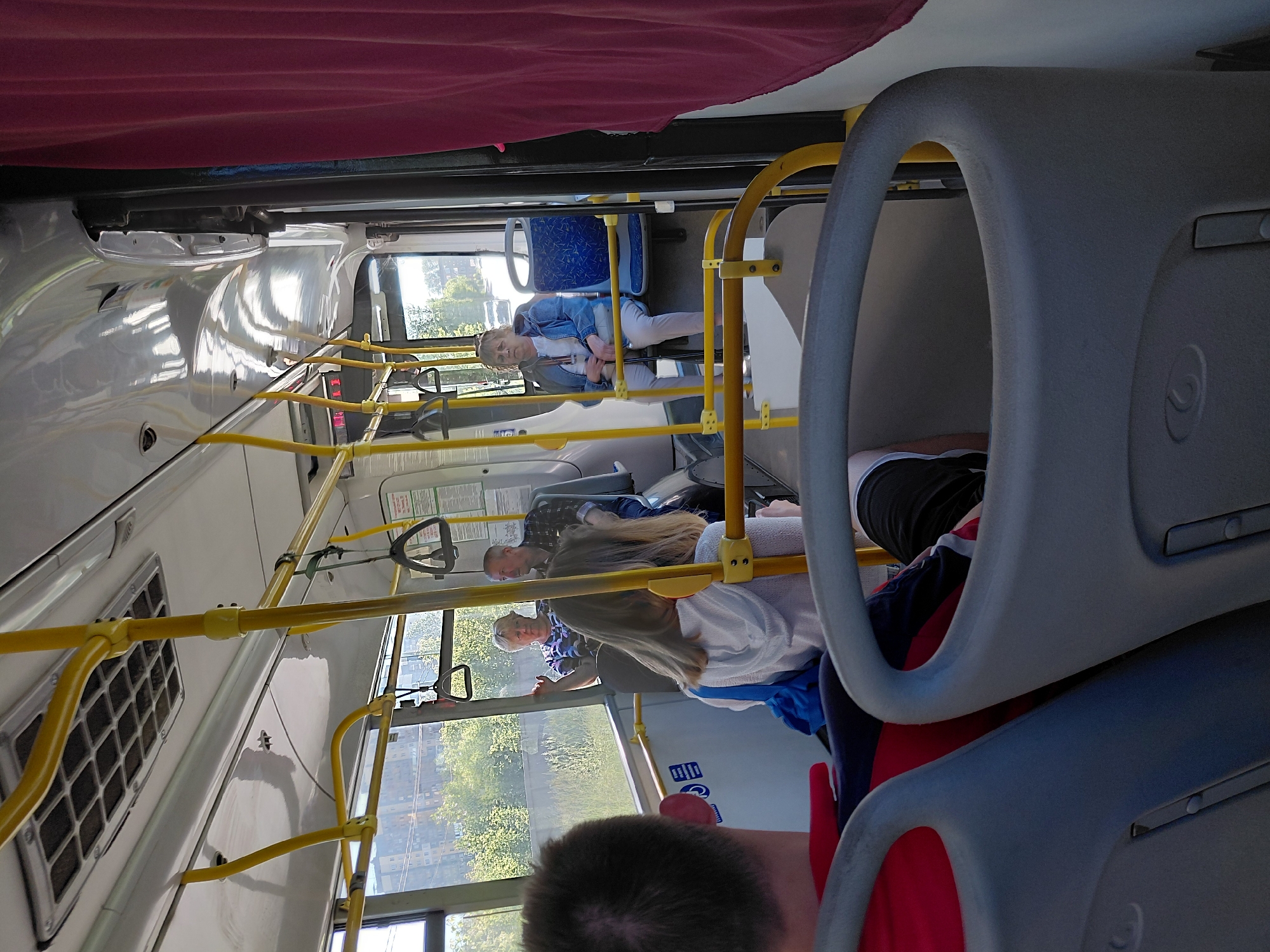 The image shows the interior of a public bus. The camera is positioned behind a person in the foreground, whose head and shoulders are visible, wearing a red shirt. Other passengers are seated throughout the bus. In the middle ground, a woman with blonde hair wearing a white hoodie is seated, facing away from the camera. Further back, two older individuals are seated near a window. On the right side of the bus, a woman in a denim jacket and white pants is seated, holding a cane. Yellow handrails and poles are visible throughout the bus, along with blue seats and a pink curtain on the far right. The environment outside the bus windows suggests an urban or suburban setting with trees and buildings visible.