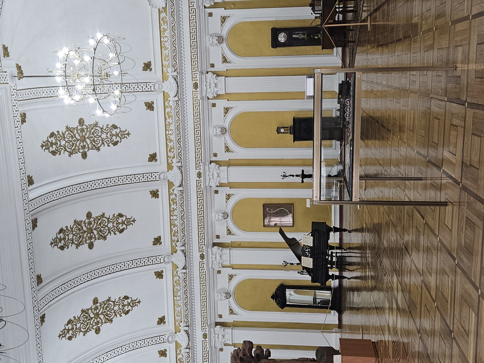 The image shows a grand hall with an ornate ceiling featuring gilded decorations and a large chandelier. The walls are painted yellow with white architectural elements, including columns and arched niches. In the center of the hall, a black grand piano with sheet music on its stand is positioned next to a tall, ornate mirror. To the right, a grandfather clock stands prominently, and further back, a display case contains a model train. A statue is partially visible on the left side of the image. The floor is made of polished wood in a geometric parquet pattern. The scene appears to be a museum or a historically preserved building. This image was taken in the National Museum of History of Moldova in Chișinău, Moldova.