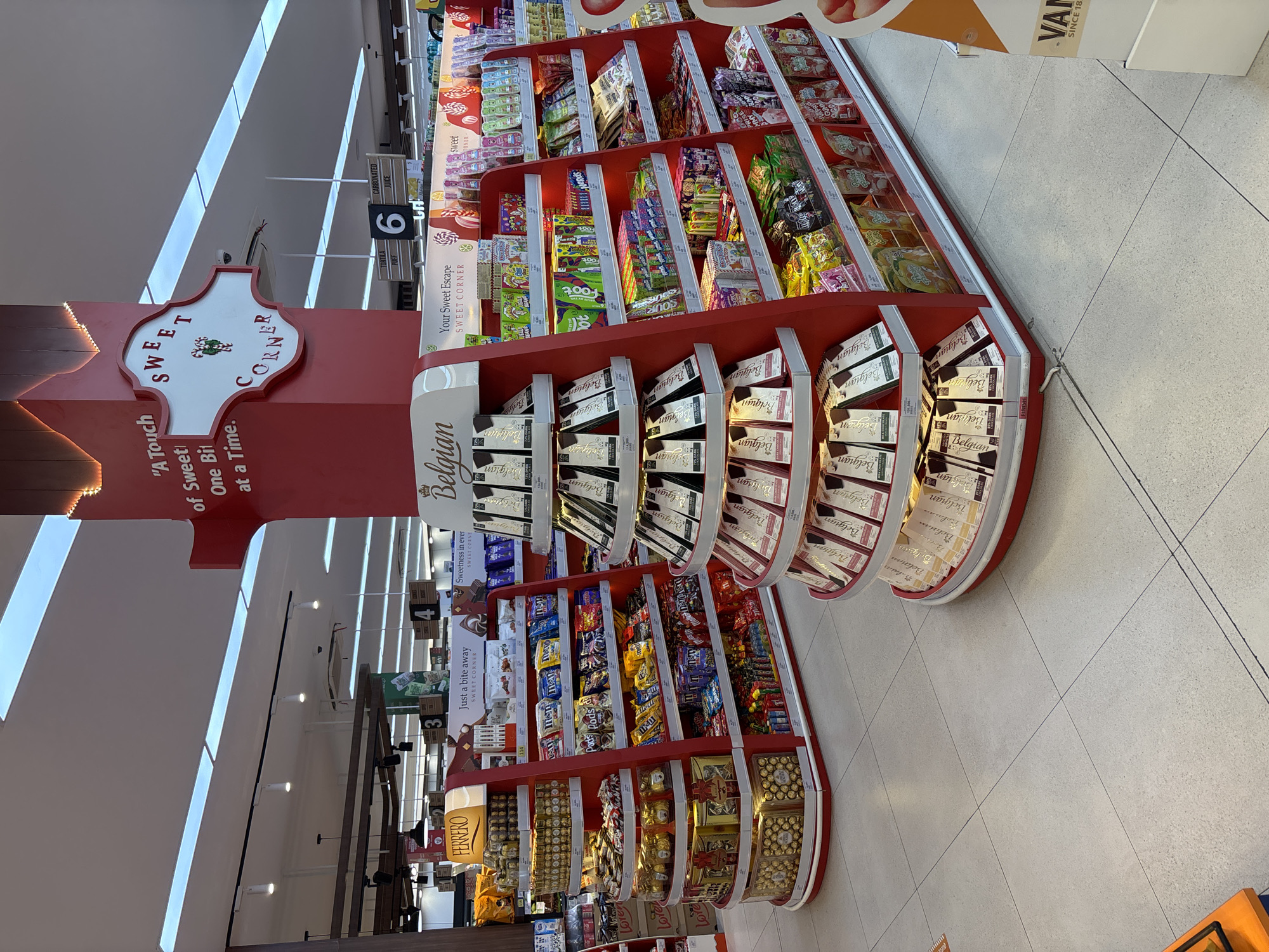 The image displays a brightly lit "Sweet Corner" section within a supermarket or grocery store in Kuta Selatan, Indonesia. The central subject is an elaborate, multi-tiered display unit, primarily red, showcasing a wide array of confectionery products. A prominent white sign in the shape of a decorative crest at the top reads "SWEET CORNER" with a small festive character, and below it, text states, "'A Touch of Sweet One Bit at a Time.'"
The shelves are heavily stocked with various candies and chocolates. The lower, curved tiers prominently feature "Belgian" branded chocolate bars, arranged in multiple rows. Higher shelves display colorful packaging for candies such as "Sour Foot," "Nerds," "Pods," and "M&M's," among other sweet treats. A dedicated section on one side is marked with a large "FERRERO" sign, holding numerous boxes of Ferrero Rocher chocolates.
Additional signs visible on the display include "Your Sweet Escape," "Sweetness in ever," and "Just a bite away," all reinforcing the theme of sweet indulgences. In the background, typical store fixtures like overhead fluorescent lights and a white-tiled floor are visible. An aisle marker showing the number "6" also lists "CARBONATED JUICE" and "TORTILLA CHIPS," indicating nearby product categories. A partially visible sign with the text "VAN SINCE 18." can be seen at the top right. The scene is well-organized and presented, typical of a retail environment, with no people visible.