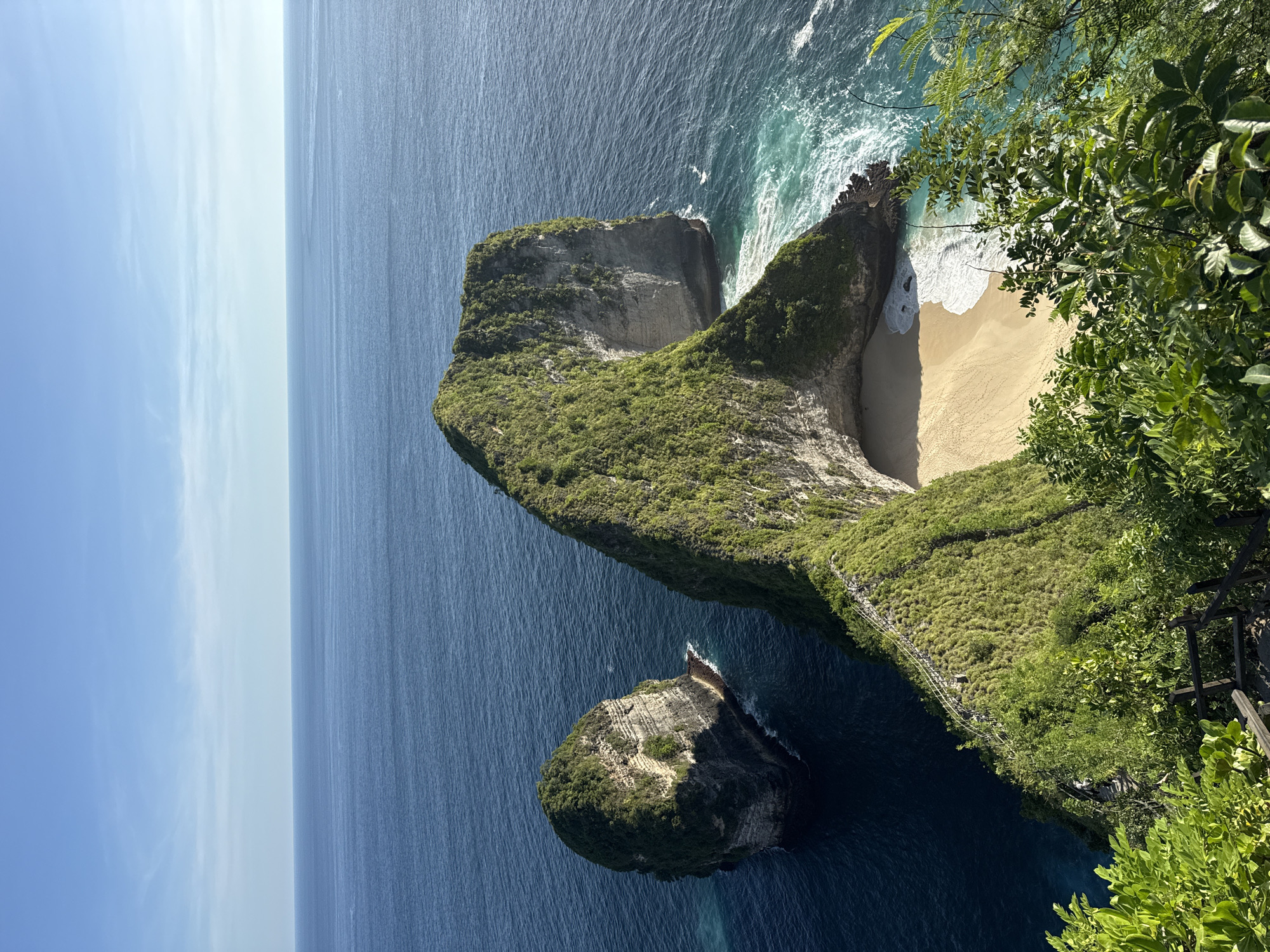 The image shows the Kelingking beach on the island of Nusa Penida, Indonesia, also known as "T-Rex beach". A promontory shaped like a Tyrannosaurus Rex is seen, with a sandy beach at its base. The cliff is covered in green vegetation. The ocean is blue and stretches to the horizon. A small islet is seen near the promontory. Waves are breaking on the shore. Trees and wooden structures can be seen in the foreground, to the right of the frame.